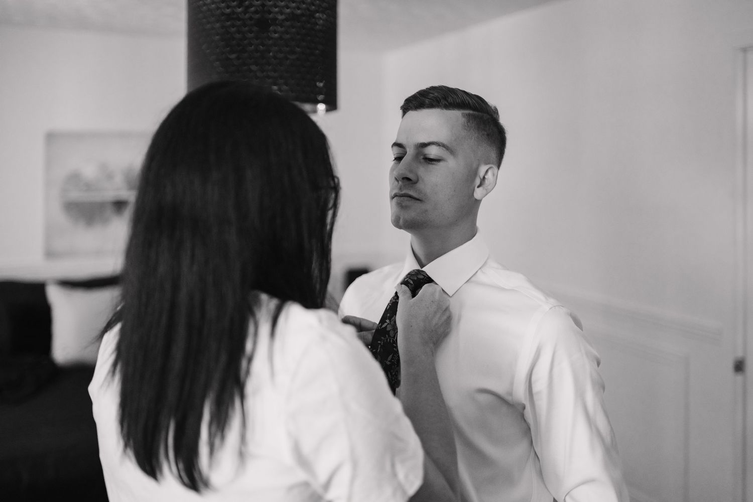 Black and white photo of a woman helping a man adjust his tie while getting dressed for a formal event.