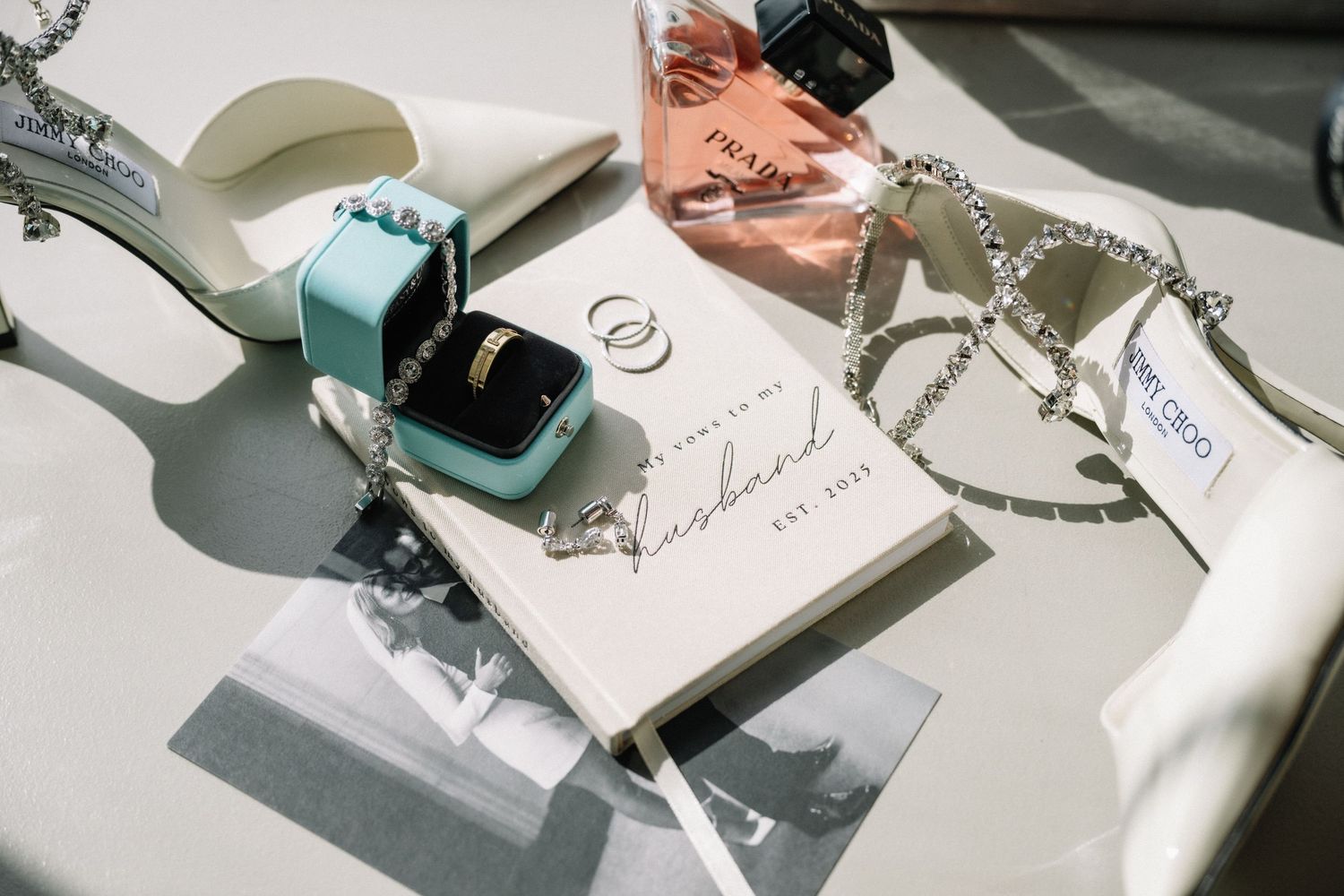 Elegant wedding flat lay with Tiffany ring box, gold band, perfume, jewelry, and personalized vow book on white surface.