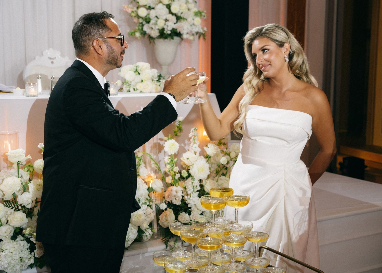 Bride and groom toasting champagne beside a pyramid of glasses at their wedding reception.