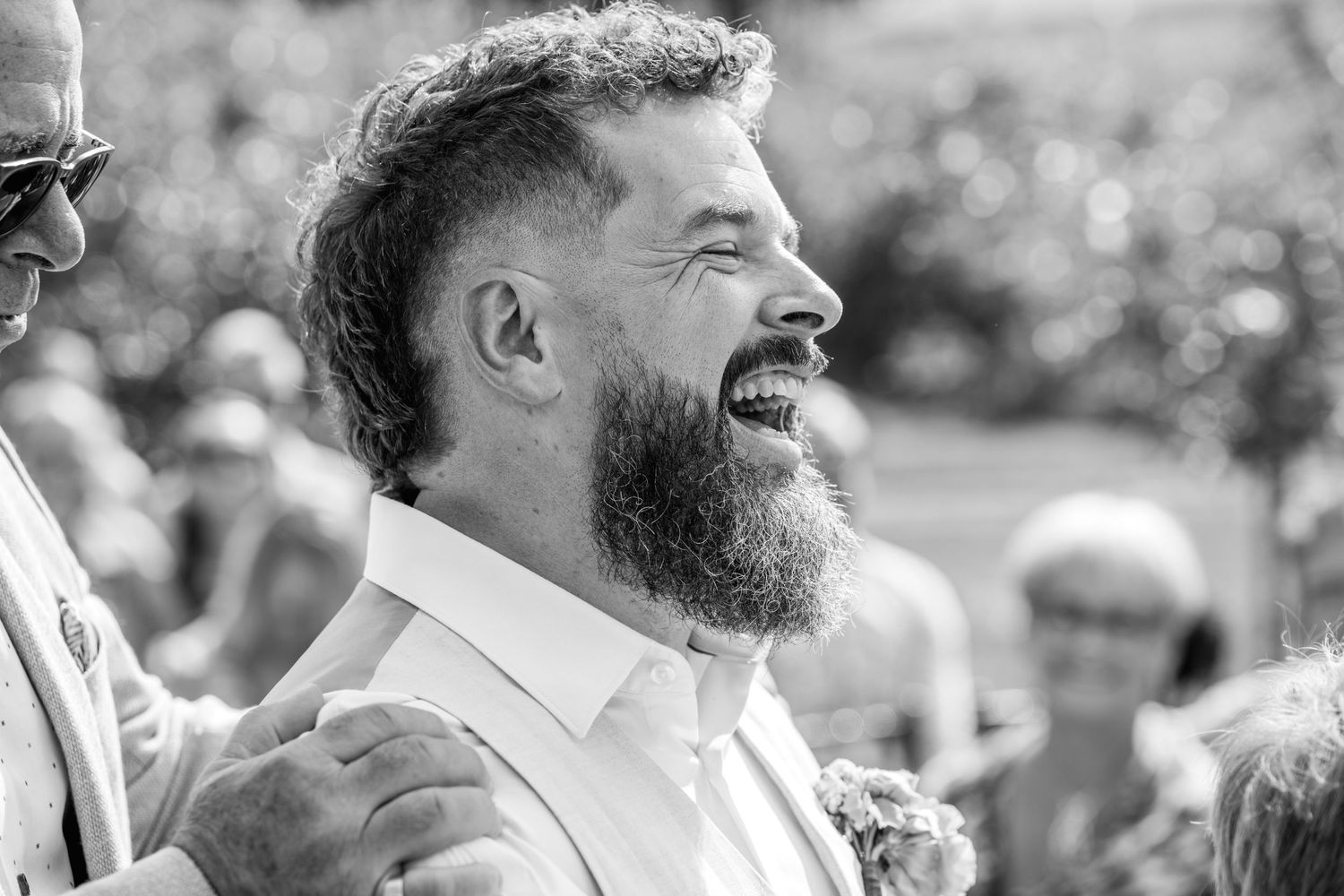 Bearded groom laughing joyfully at outdoor wedding ceremony in black and white photo.