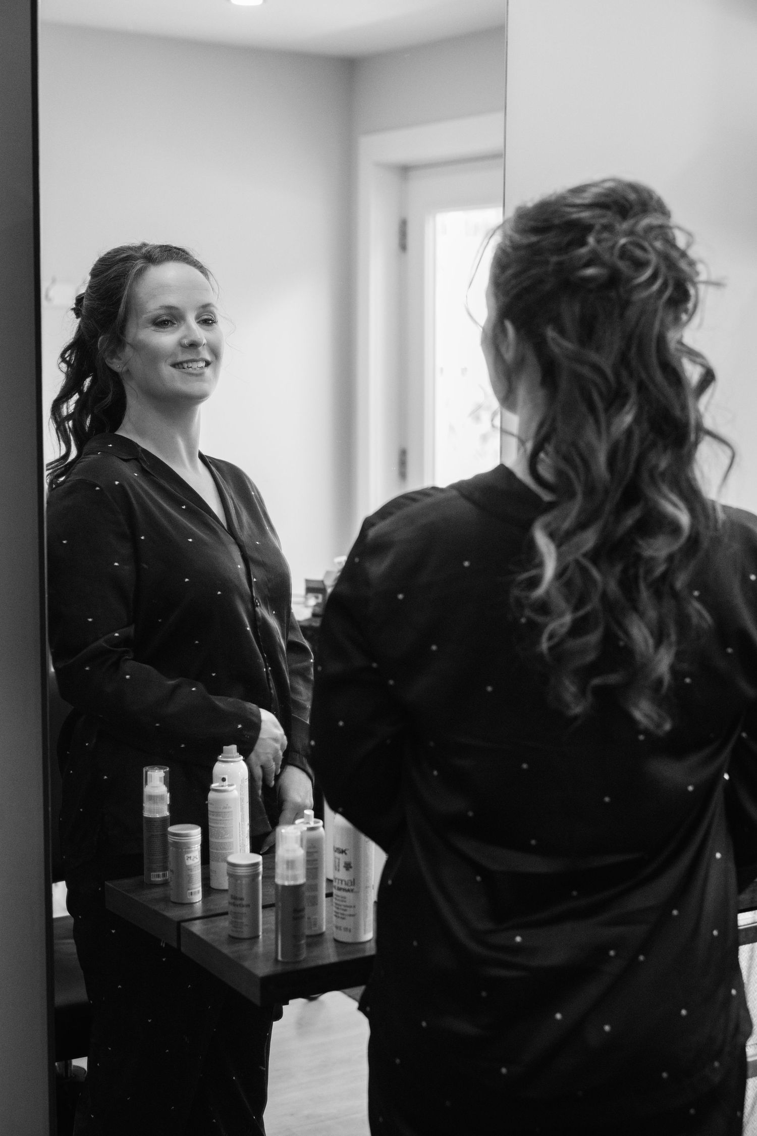 Bride smiling at her reflection in a mirror while getting ready, wearing a black robe with curled hair.