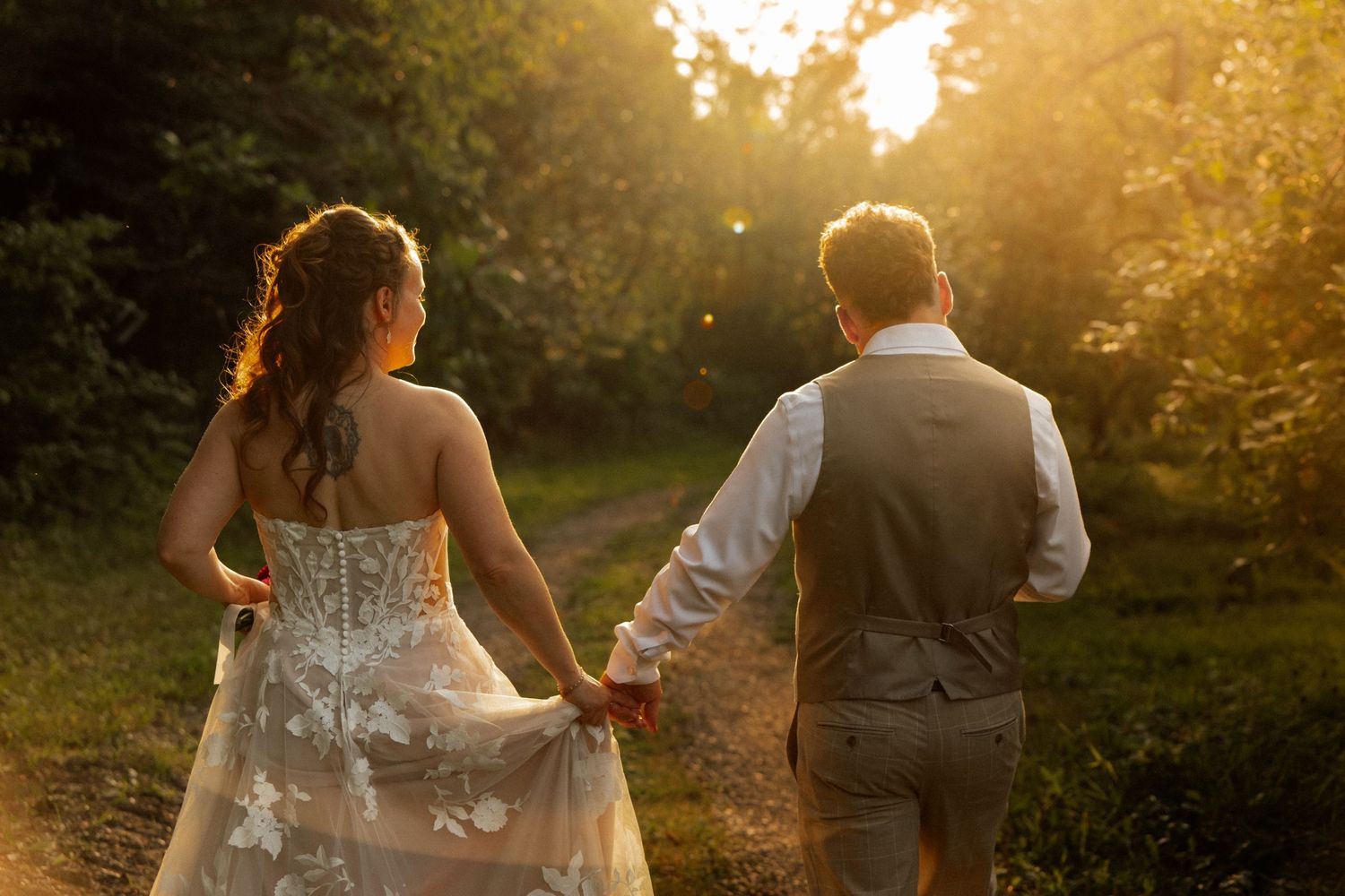 A bride and groom walk hand in hand through a sunlit forest path, captured from behind in warm golden hour light.