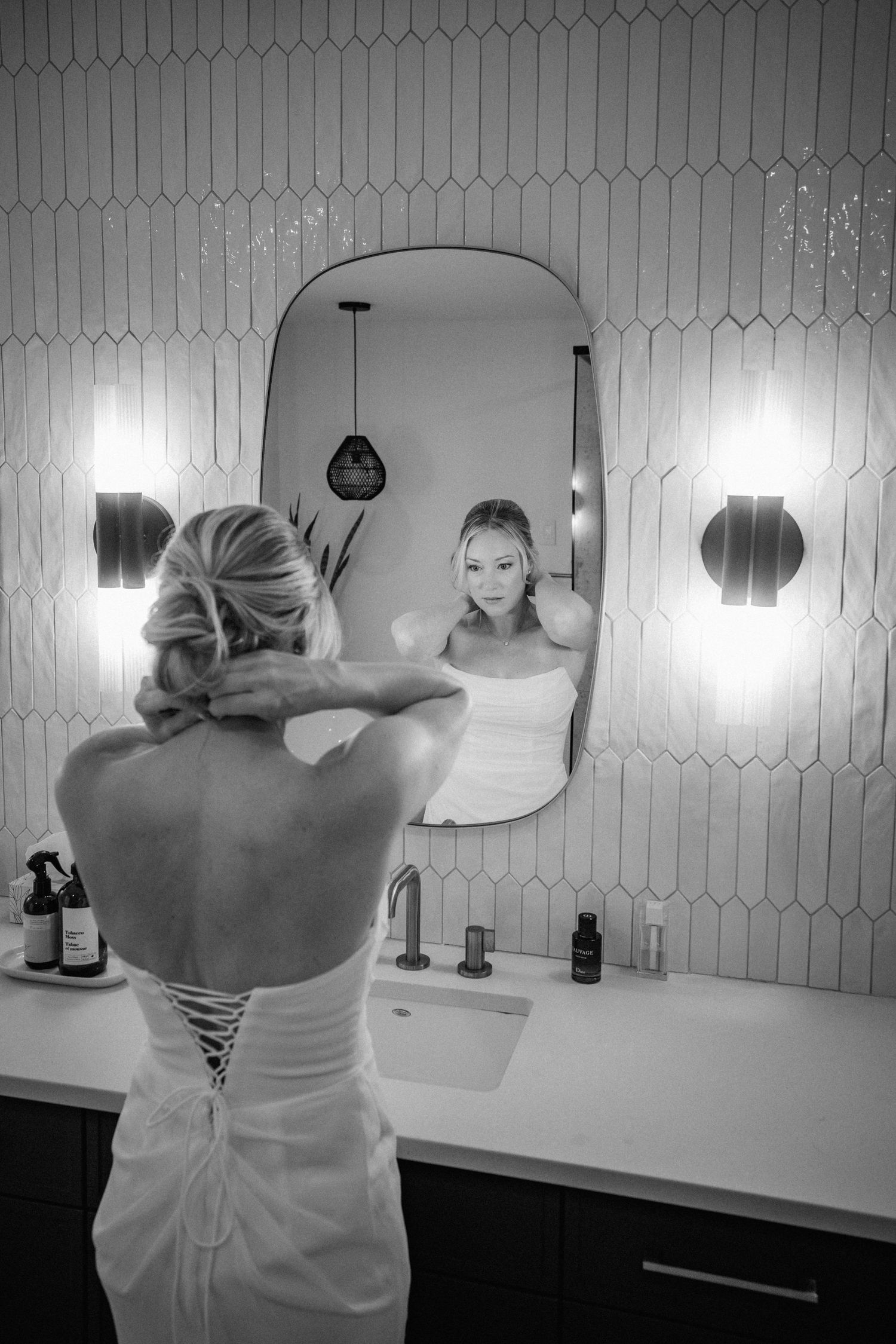 Black and white photo of a bride in a lace-back gown adjusting her updo while gazing at her reflection in a bathroom mirror.