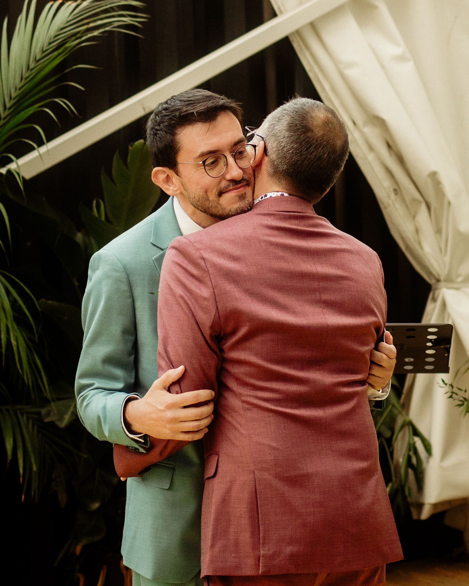 Two grooms in mint and mauve suits share an emotional embrace during their wedding ceremony.