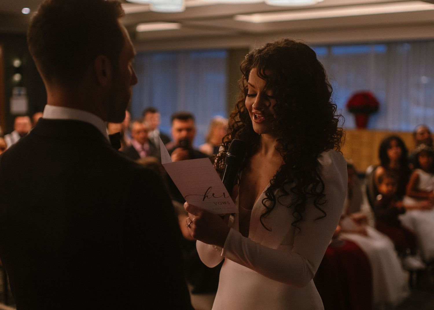 Bride smiling while reading wedding vows to groom during indoor ceremony with guests seated in background.