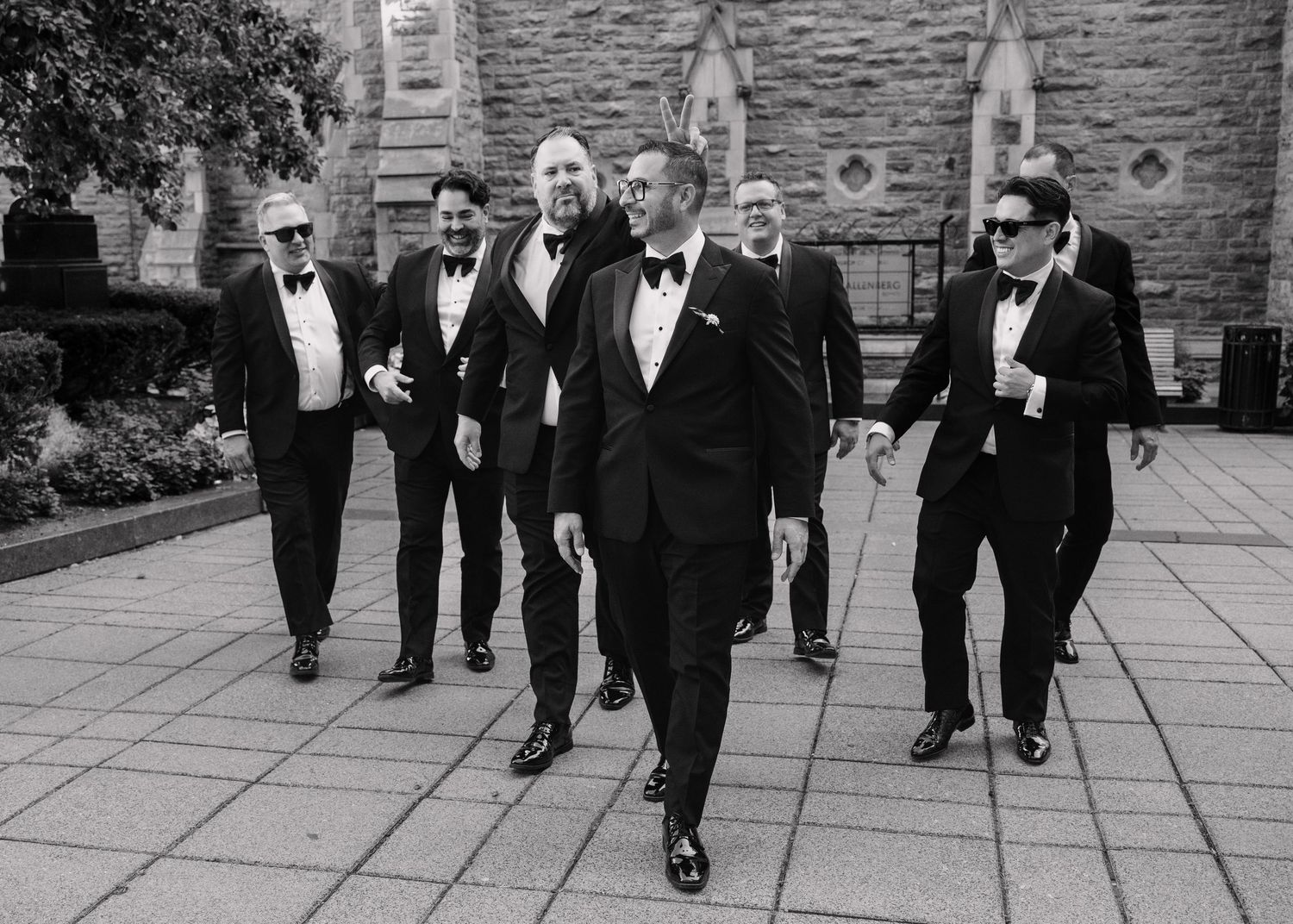 Groom and groomsmen in black tuxedos walking confidently outside stone church.