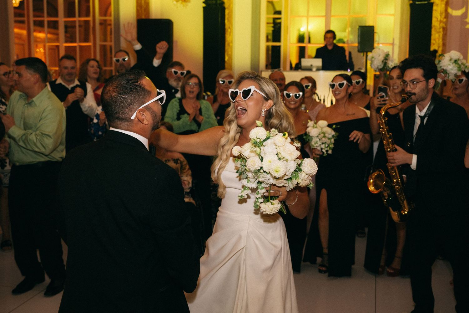Bride in white gown and sunglasses dances with groom at lively wedding reception with live saxophone music.