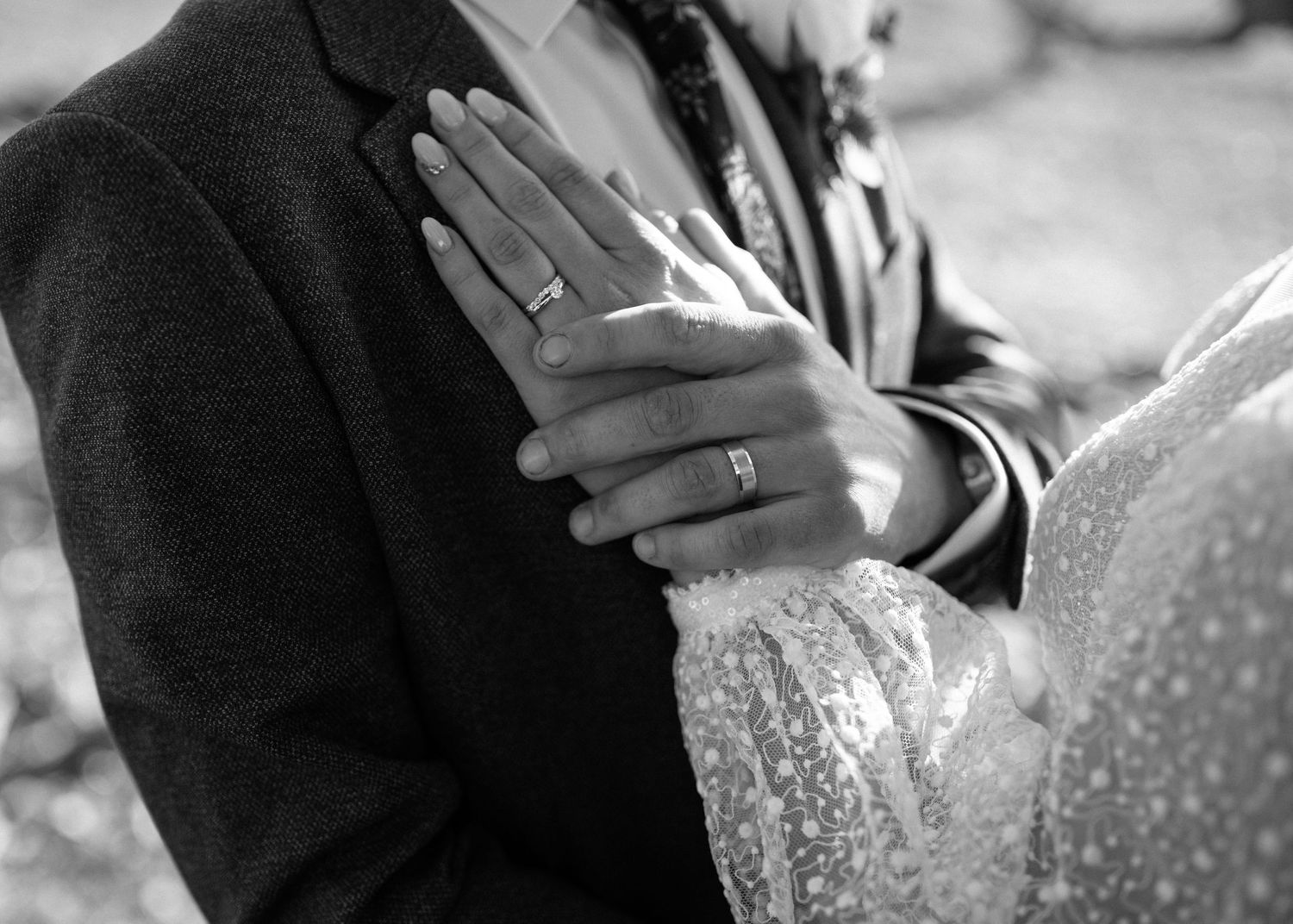 Black and white close-up of bride and groom hands showing wedding rings against formal attire.