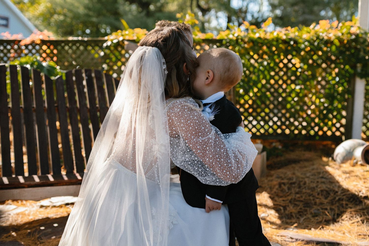 Bride in white gown and veil holds a young child in a tuxedo at an outdoor wedding.