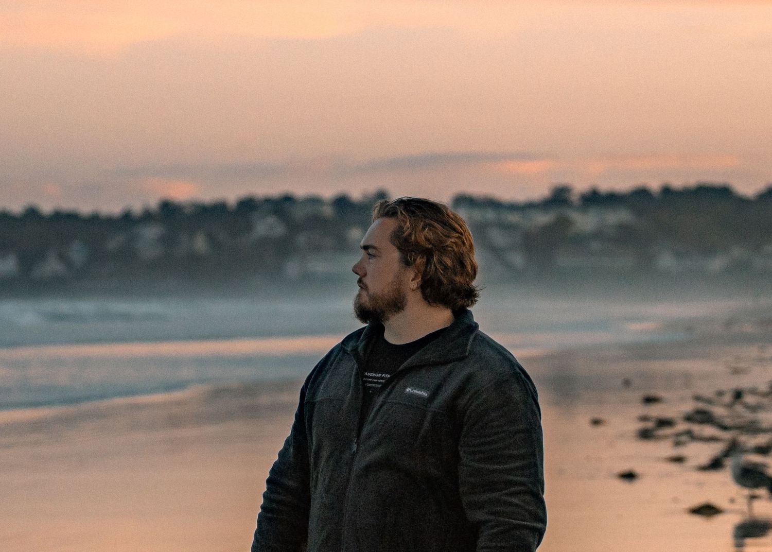 Bearded man in black jacket standing on a beach at sunset with a coastal town in the background.