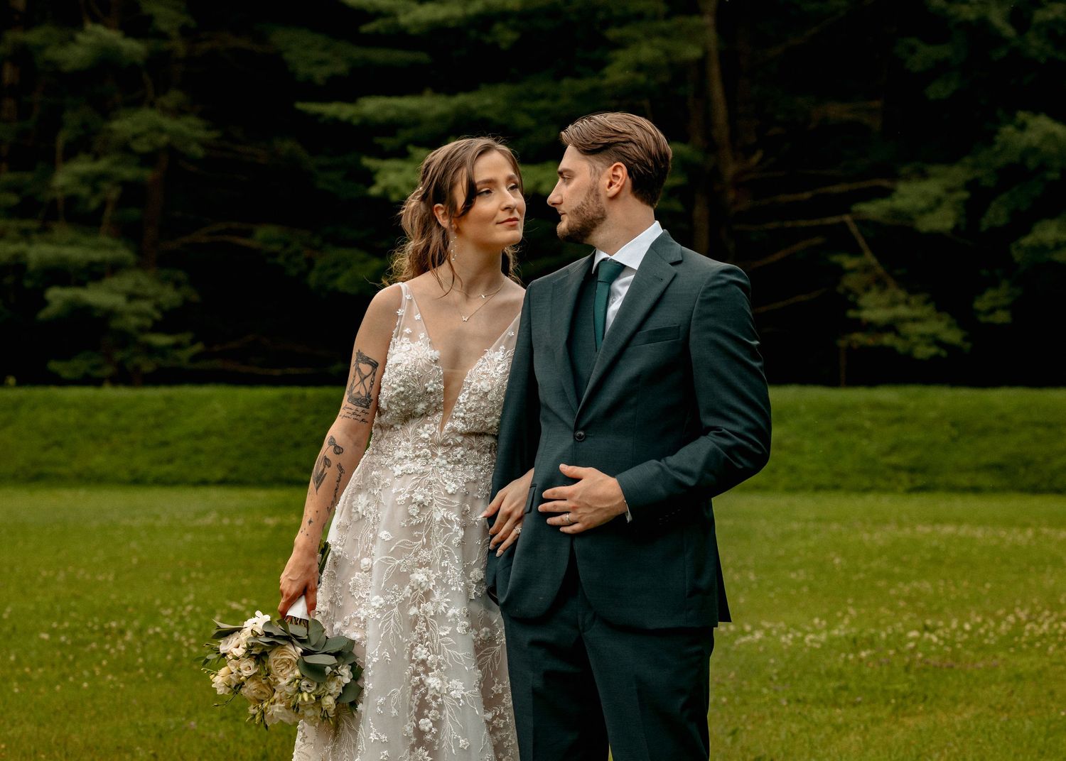 Bride and groom share a romantic moment on green lawn surrounded by tall trees.