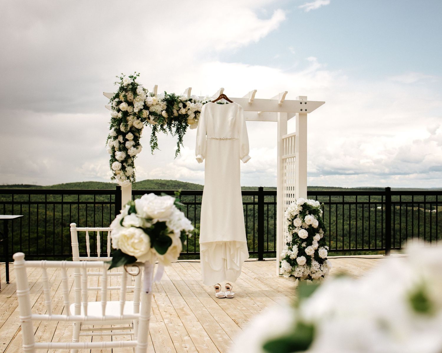 Elegant outdoor wedding ceremony setup with white floral arch, bridal gown hanging, and white chairs on a deck overlooking scenic hills.