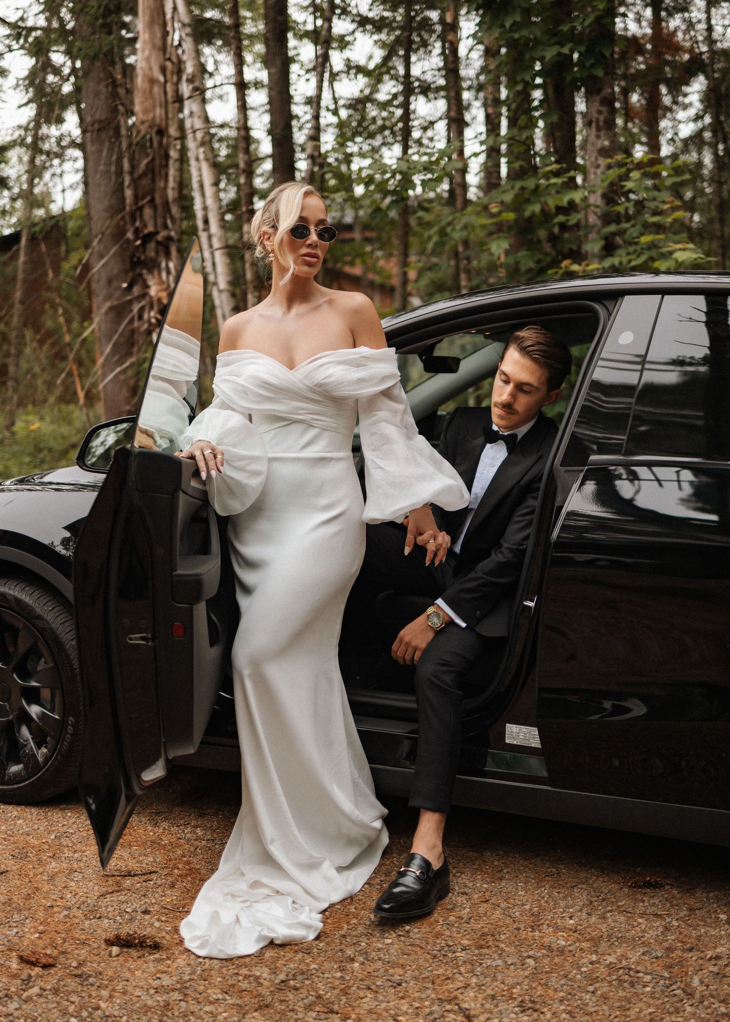 Bride in white off-shoulder gown and groom in black tuxedo posing by a black car in a forest setting.