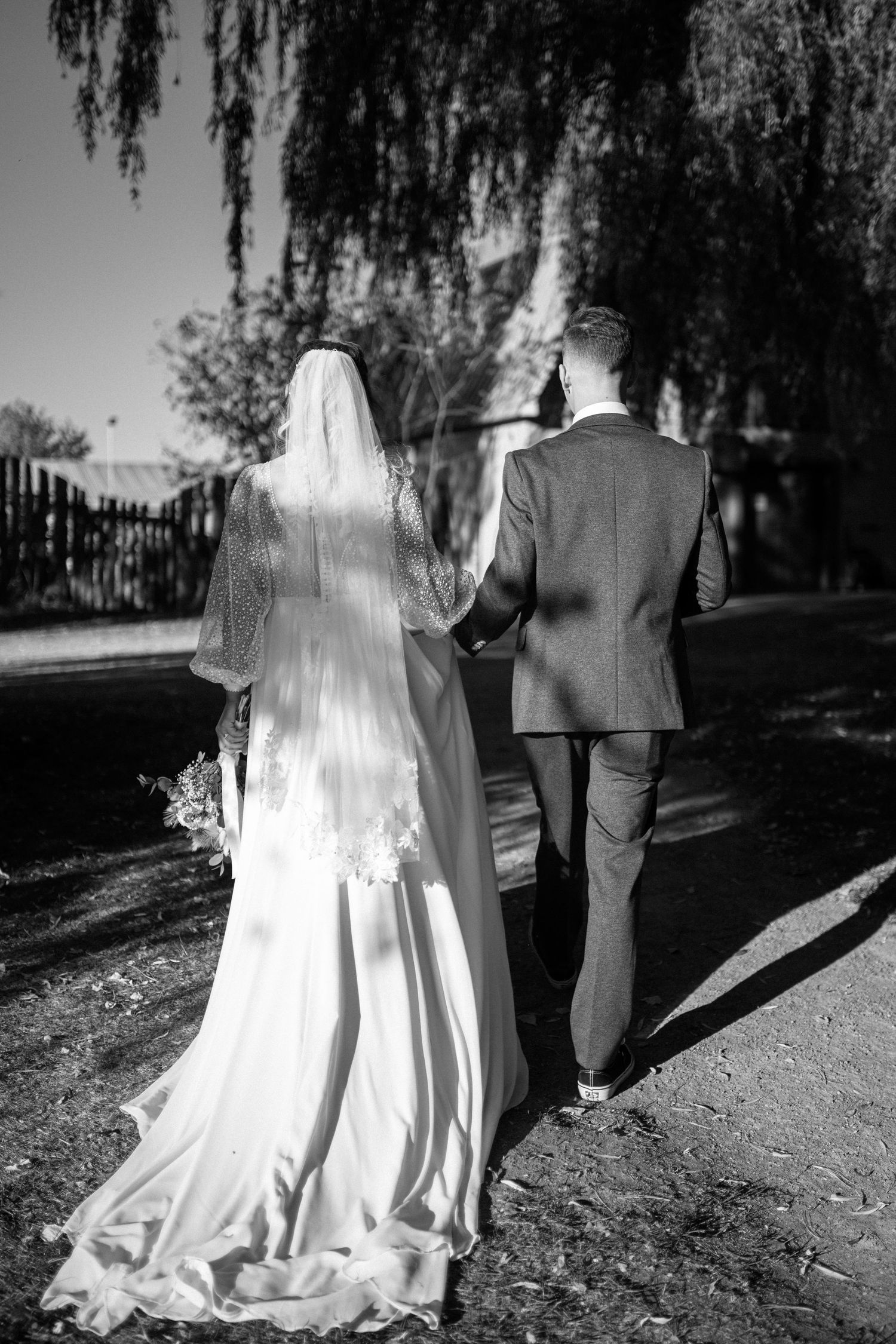 Bride and groom walking hand in hand down a sunny path in a black and white photo.