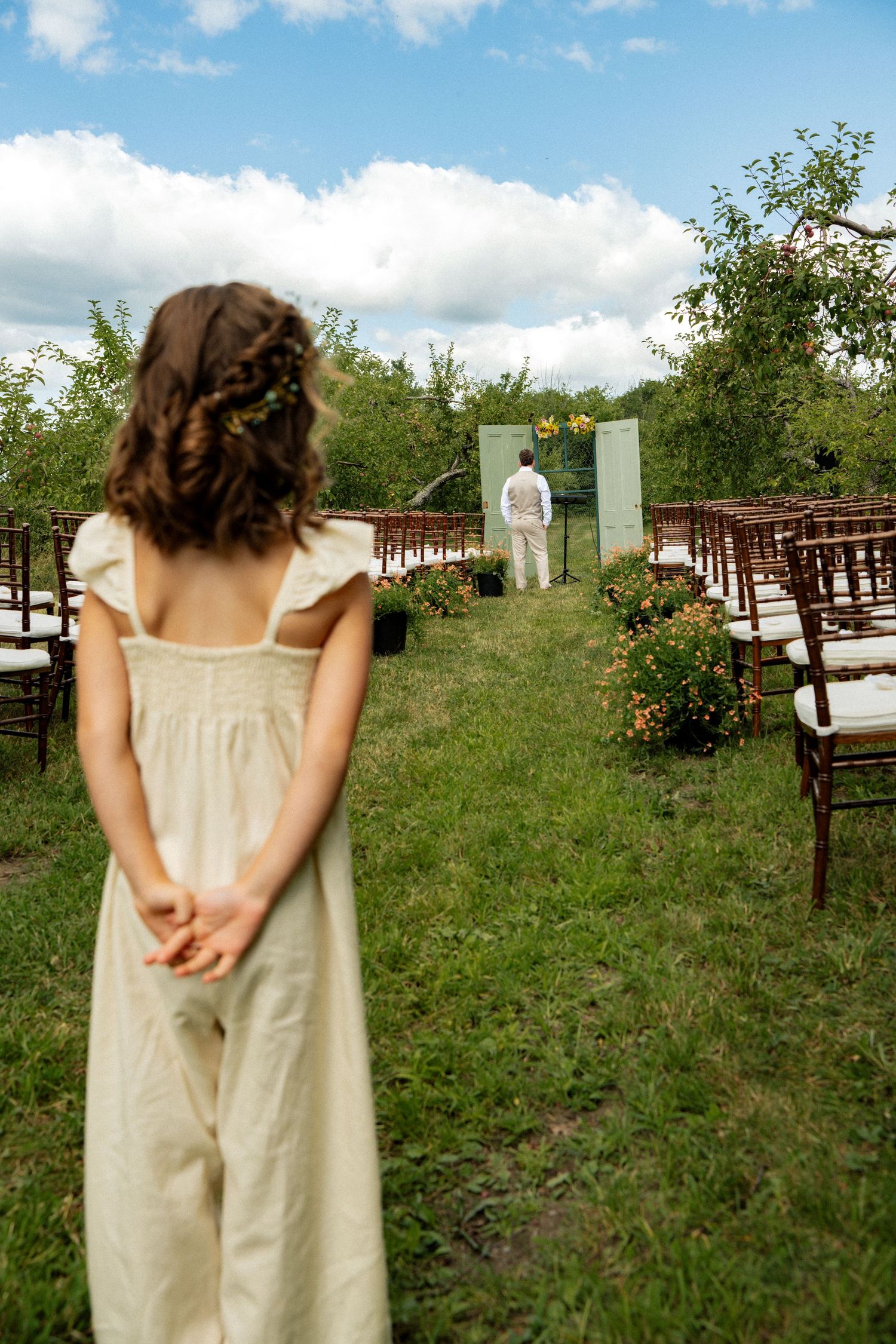 A young girl in a white dress with a floral crown stands at the entrance of an outdoor wedding ceremony.
