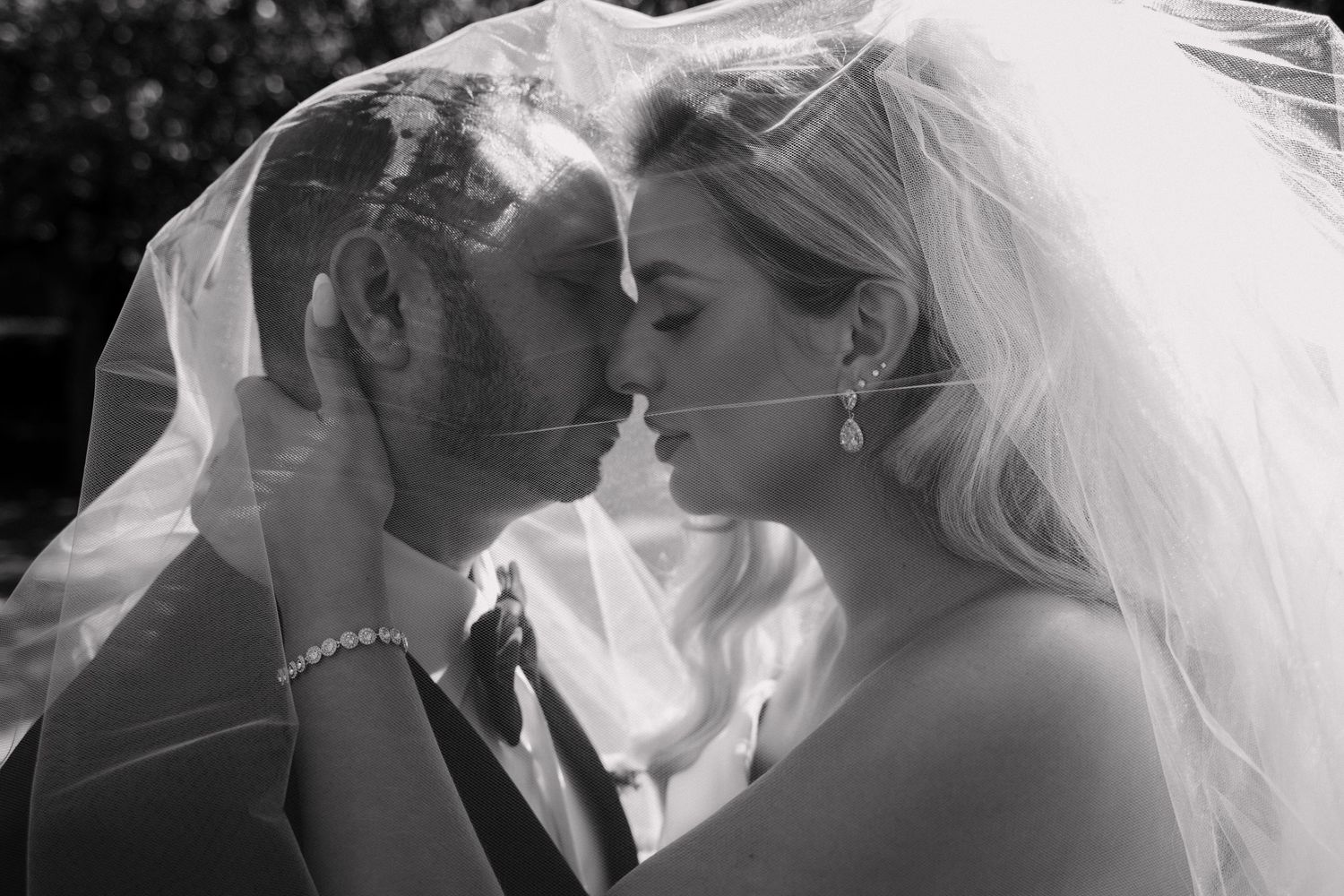 Black and white romantic photo of bride and groom sharing intimate moment under flowing wedding veil.