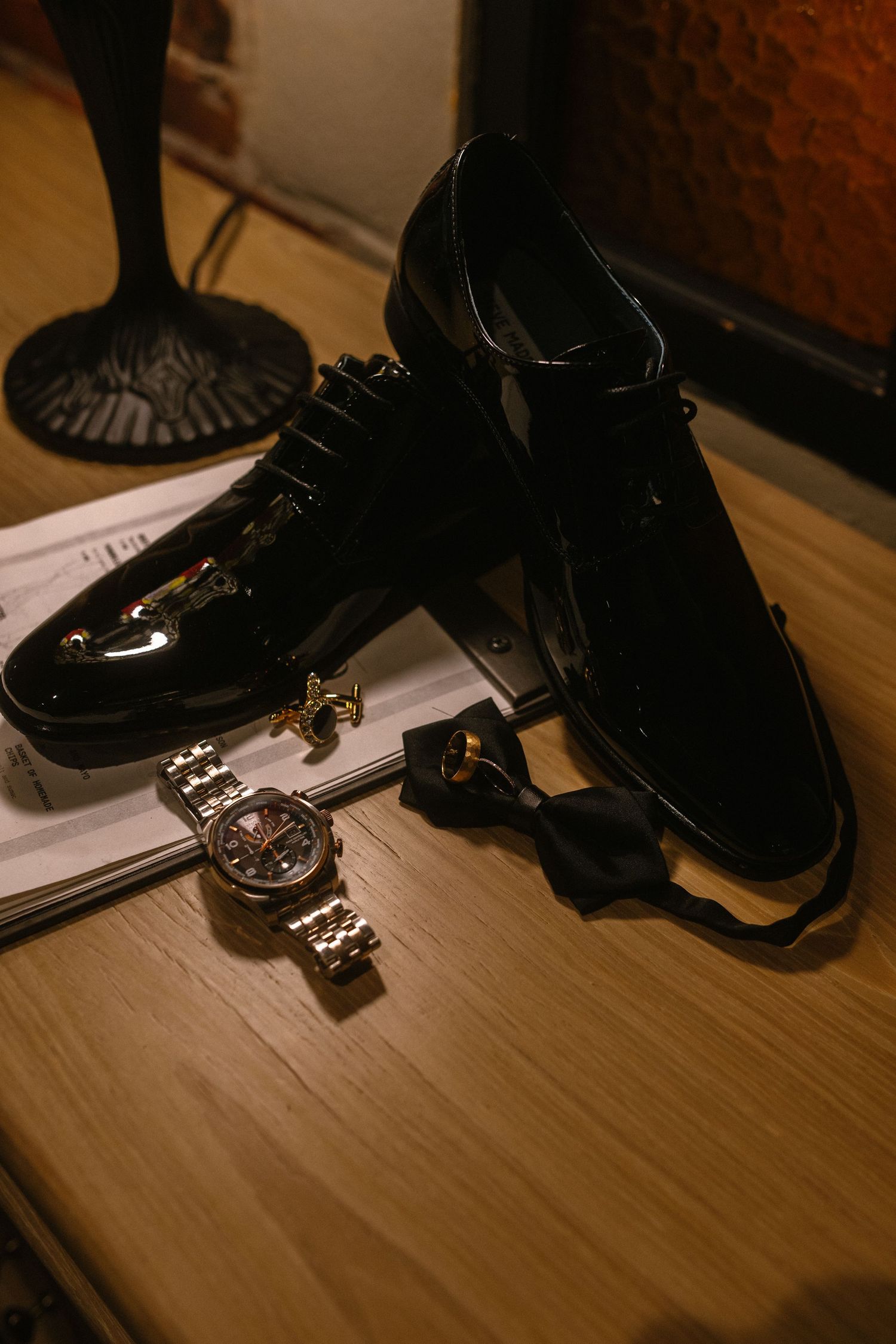 Black patent leather dress shoes with gold cufflinks, bow tie, and luxury watch on wooden surface.