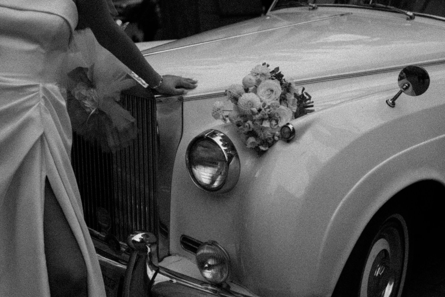 Black and white close-up of a bride beside a vintage Rolls-Royce decorated with a floral bouquet.