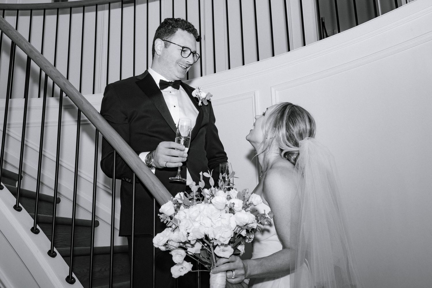 Bride holding bouquet and groom with champagne share a smile on elegant staircase in black and white.