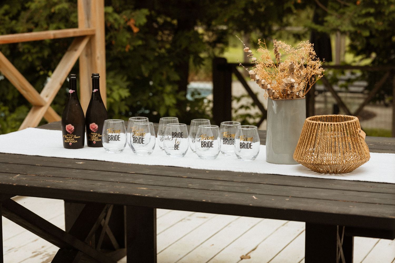 Outdoor wooden table set with wine glasses and dark bottles beside a wicker basket and dried floral arrangement on a deck.