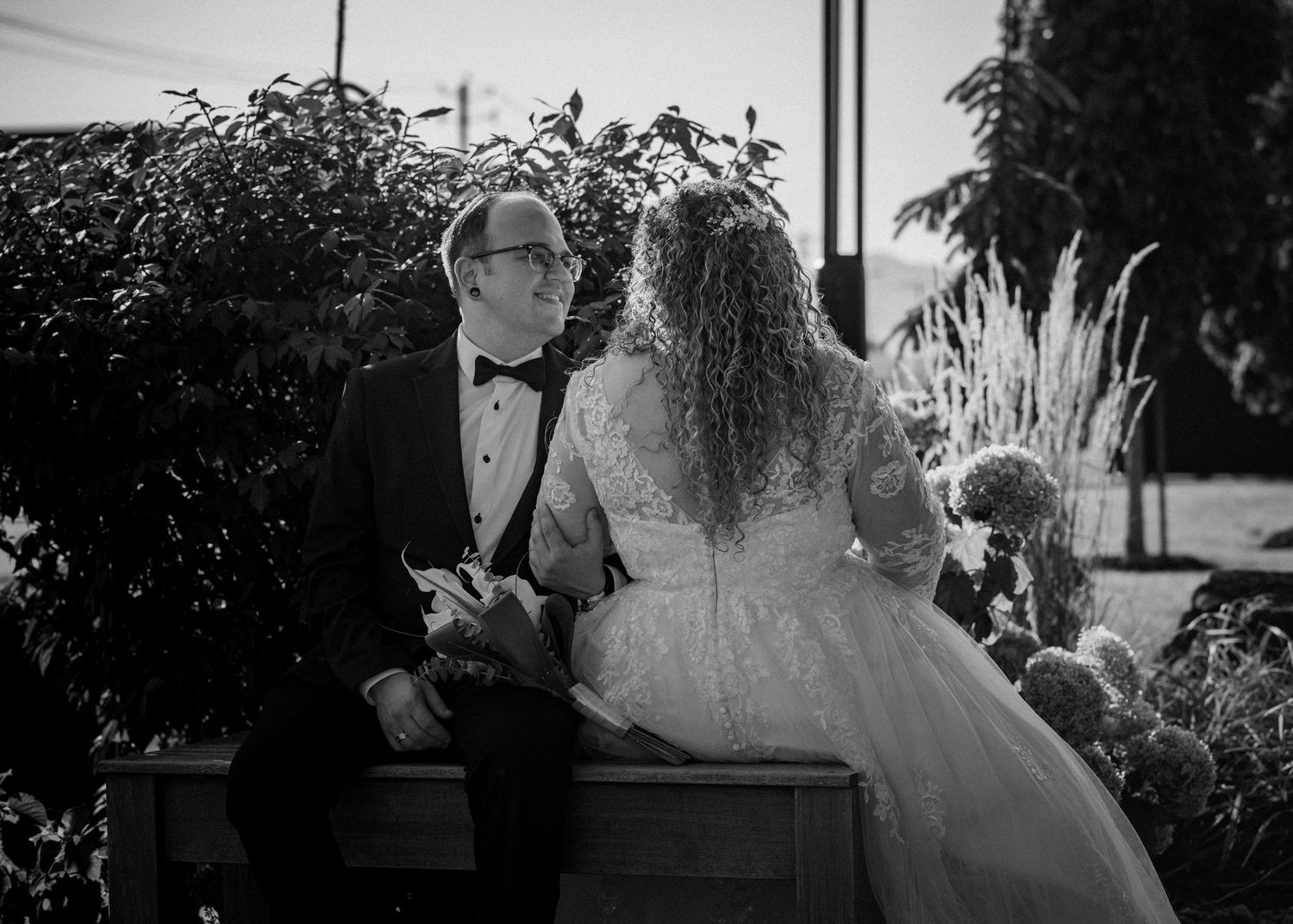 Black and white photo of a bride and groom sitting together outdoors, surrounded by lush greenery on their wedding day.