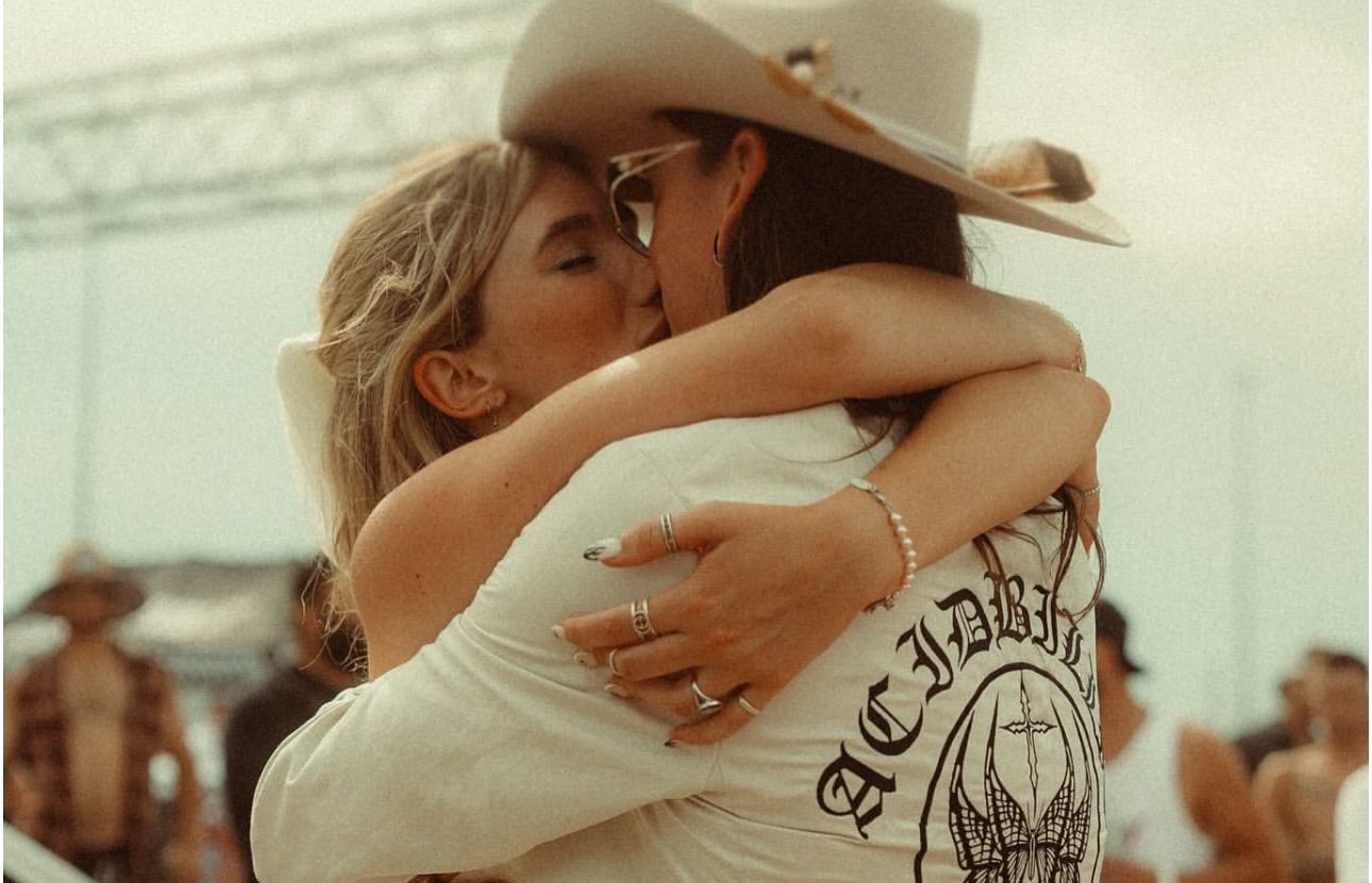 Two women share an emotional hug at an outdoor country music festival, one wearing a cowboy hat.