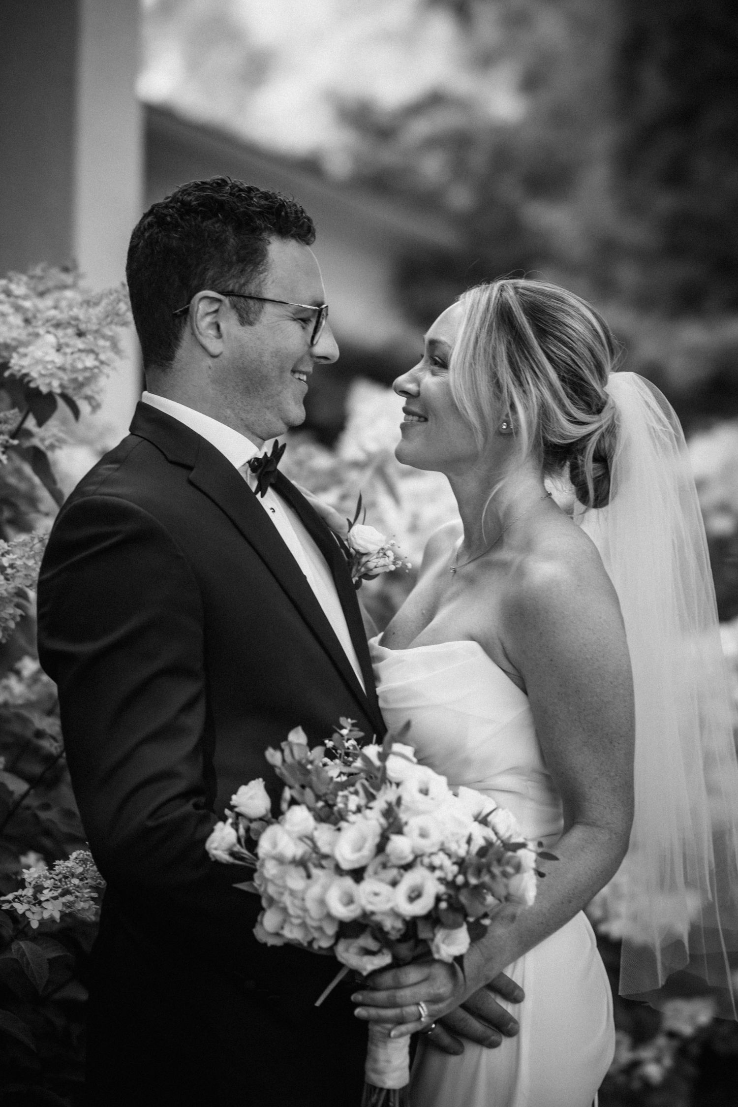 Black and white photo of smiling bride and groom holding bouquet in garden.