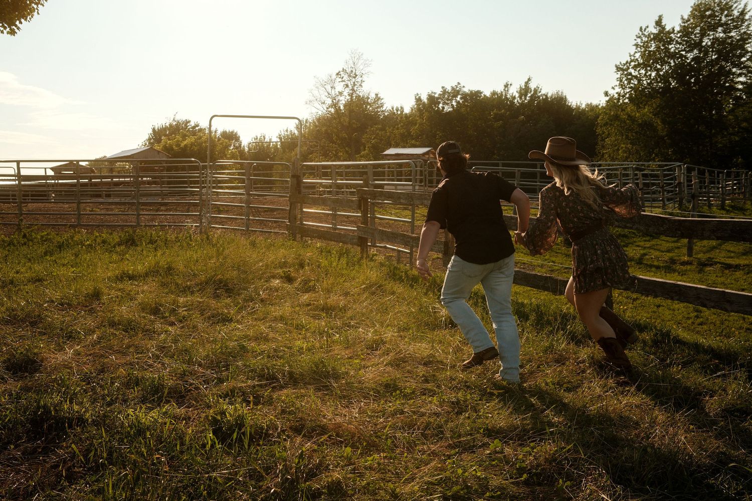 Couple running together through sunlit green pasture near ranch fence during golden hour sunset.