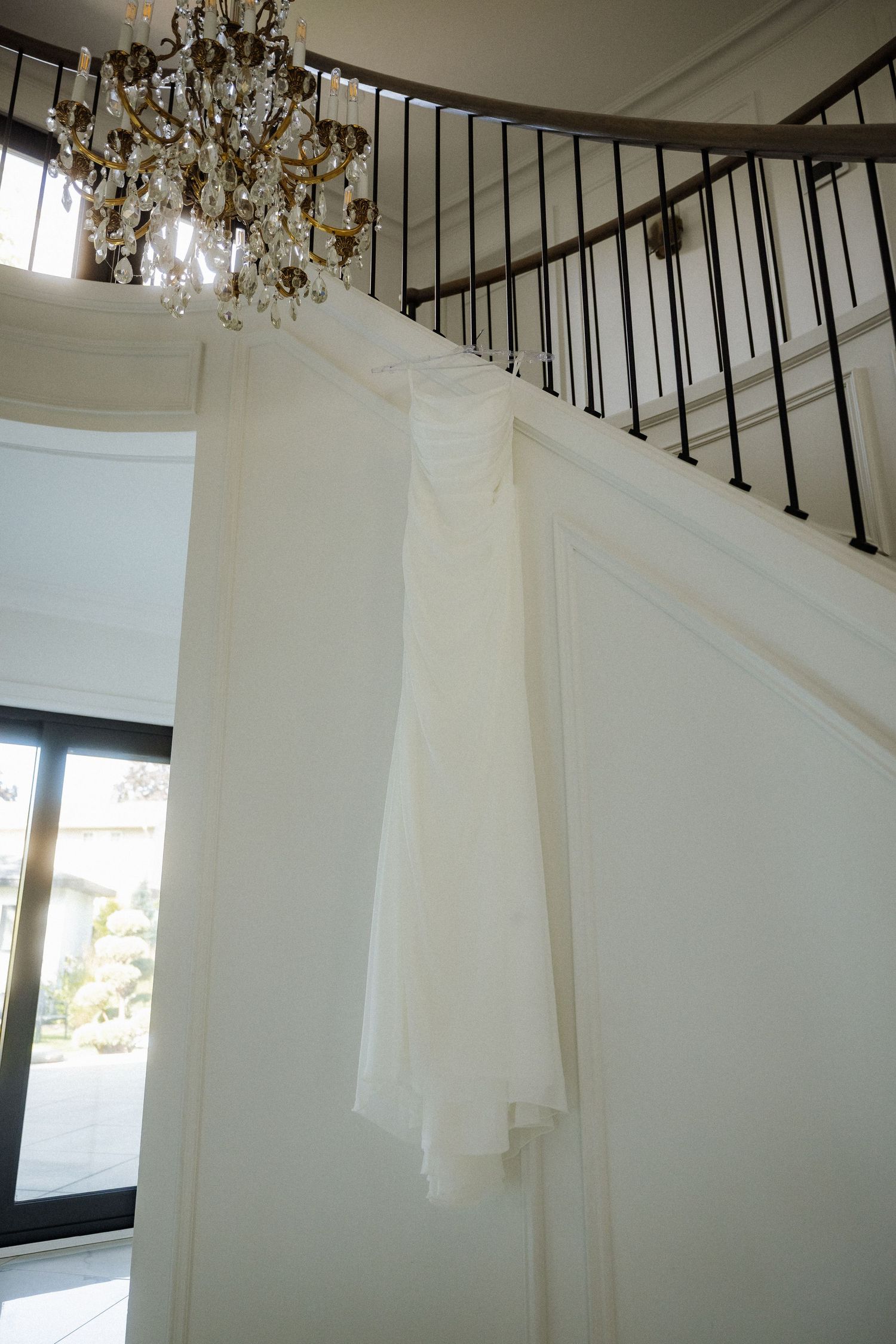 Elegant white wedding dress hanging on a curved staircase beneath a crystal chandelier in a bright white interior.