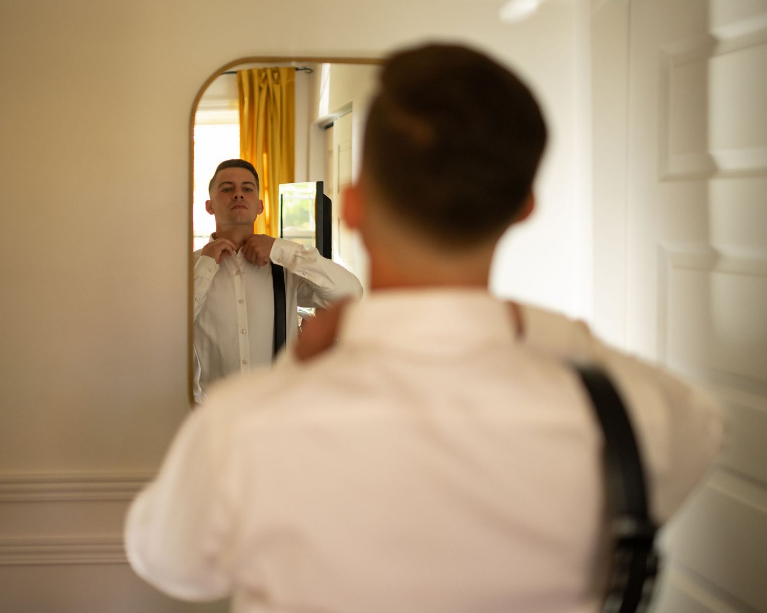 Groom adjusting his collar in a gold-framed mirror while wearing a white dress shirt and suspenders.