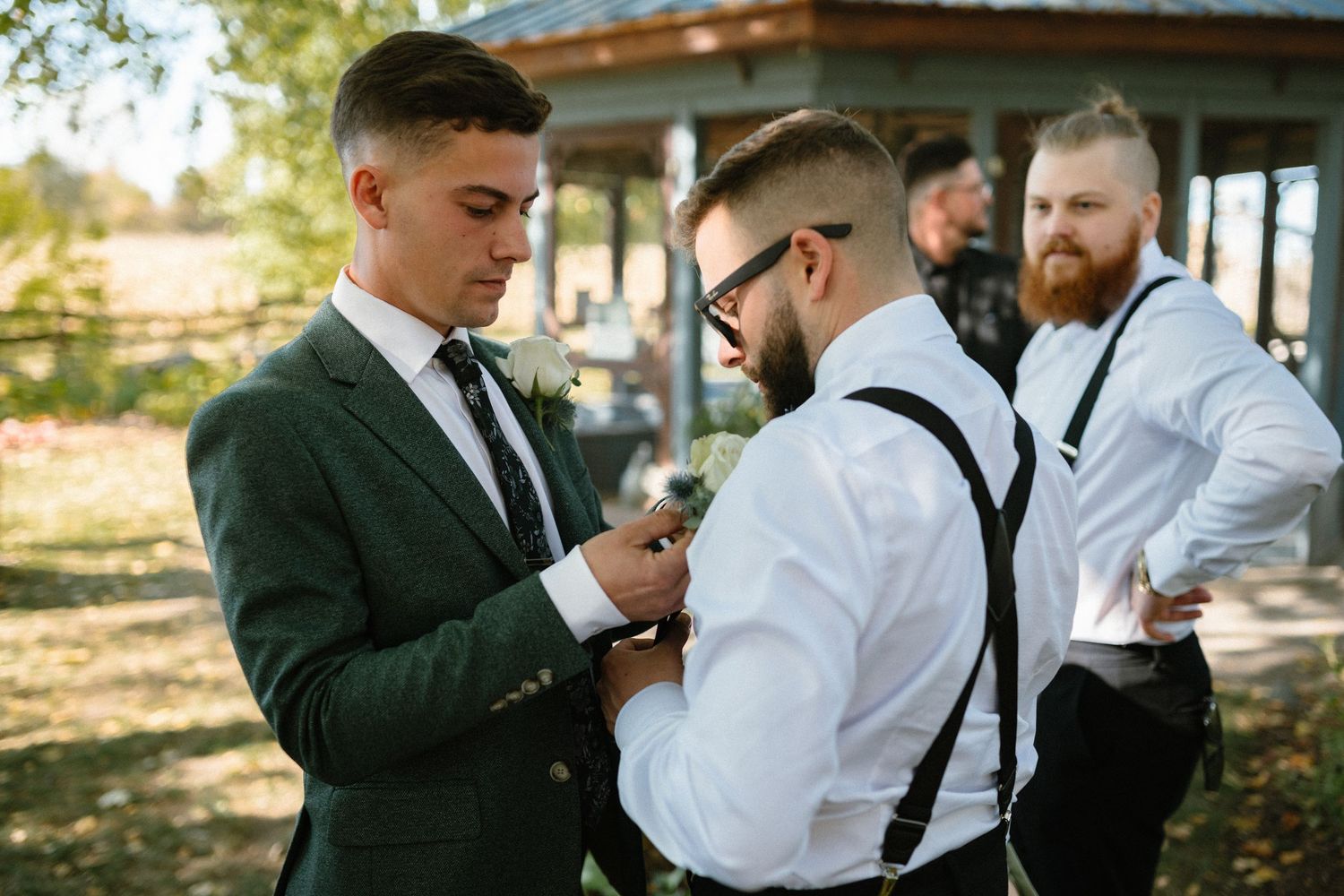 Groomsmen in green suits and suspenders helping each other with boutonnieres before a wedding.