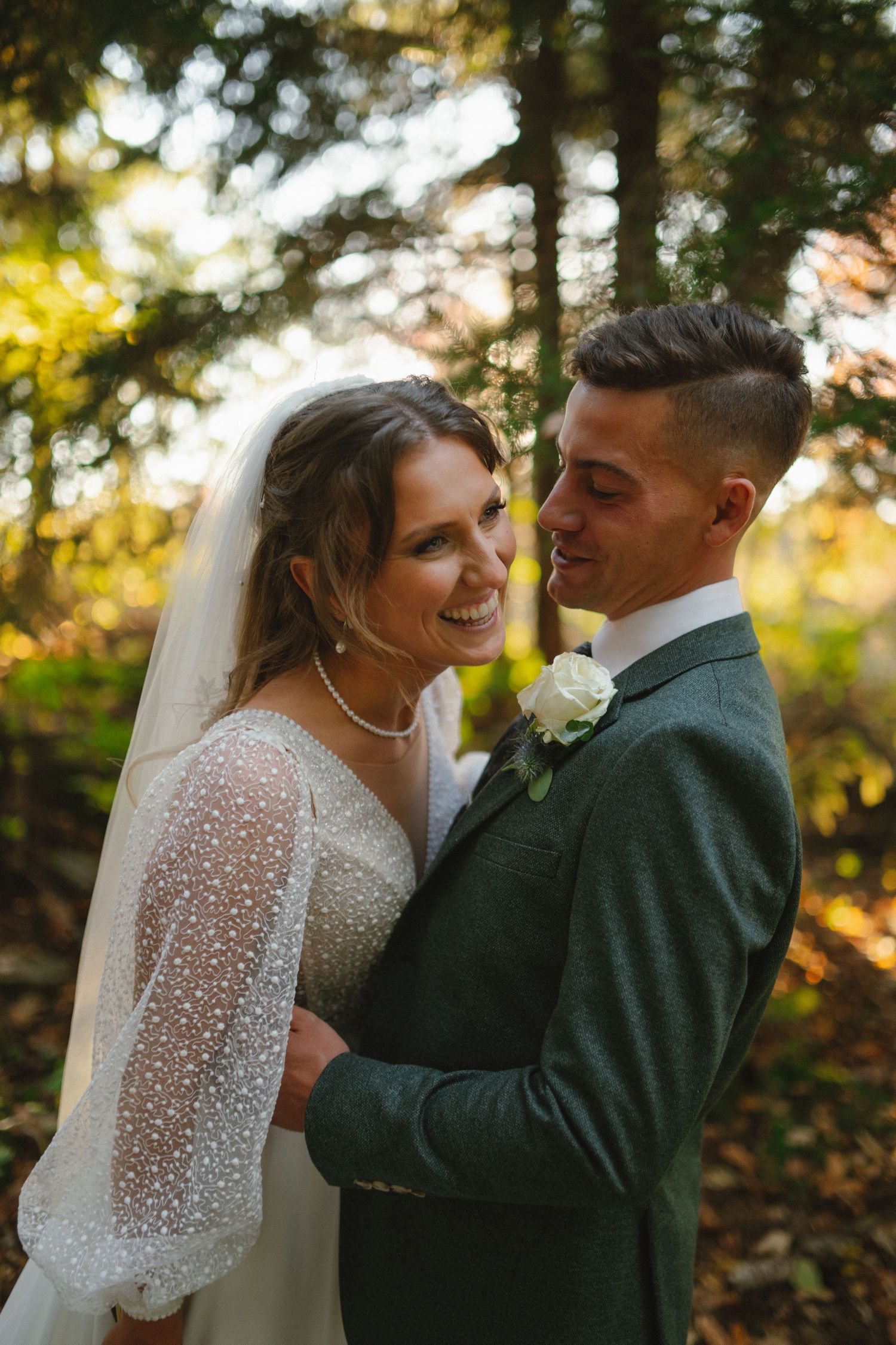 A smiling bride in a pearl-detailed white gown embraces her groom in a green suit among autumn forest trees.