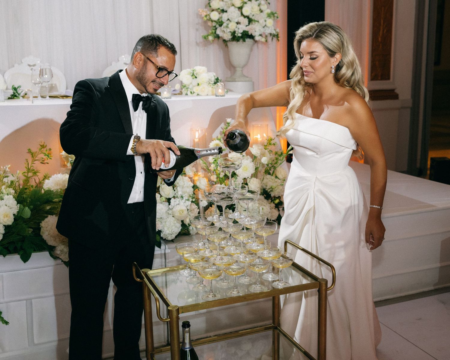 Bride and groom pour champagne tower at elegant wedding reception with white floral arrangements.