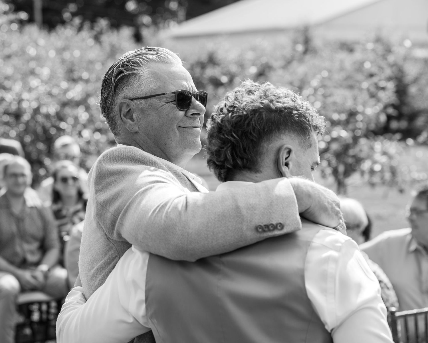 Two men sharing a warm embrace at an outdoor wedding ceremony in black and white.