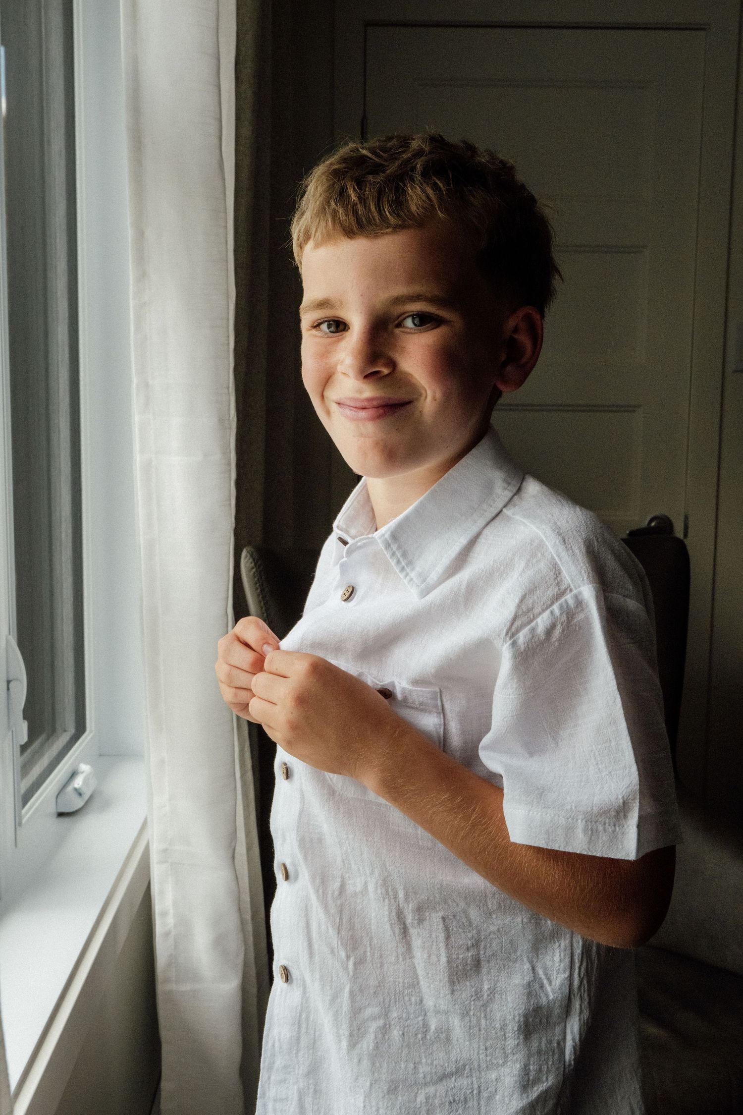 Smiling young boy in white linen shirt standing by a sunlit window.