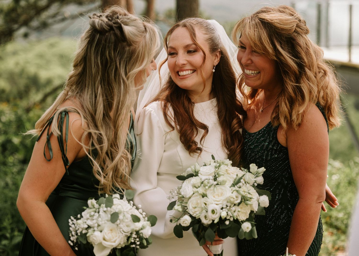 Bride in white gown and veil smiles with two bridesmaids in dark green dresses holding white floral bouquets.