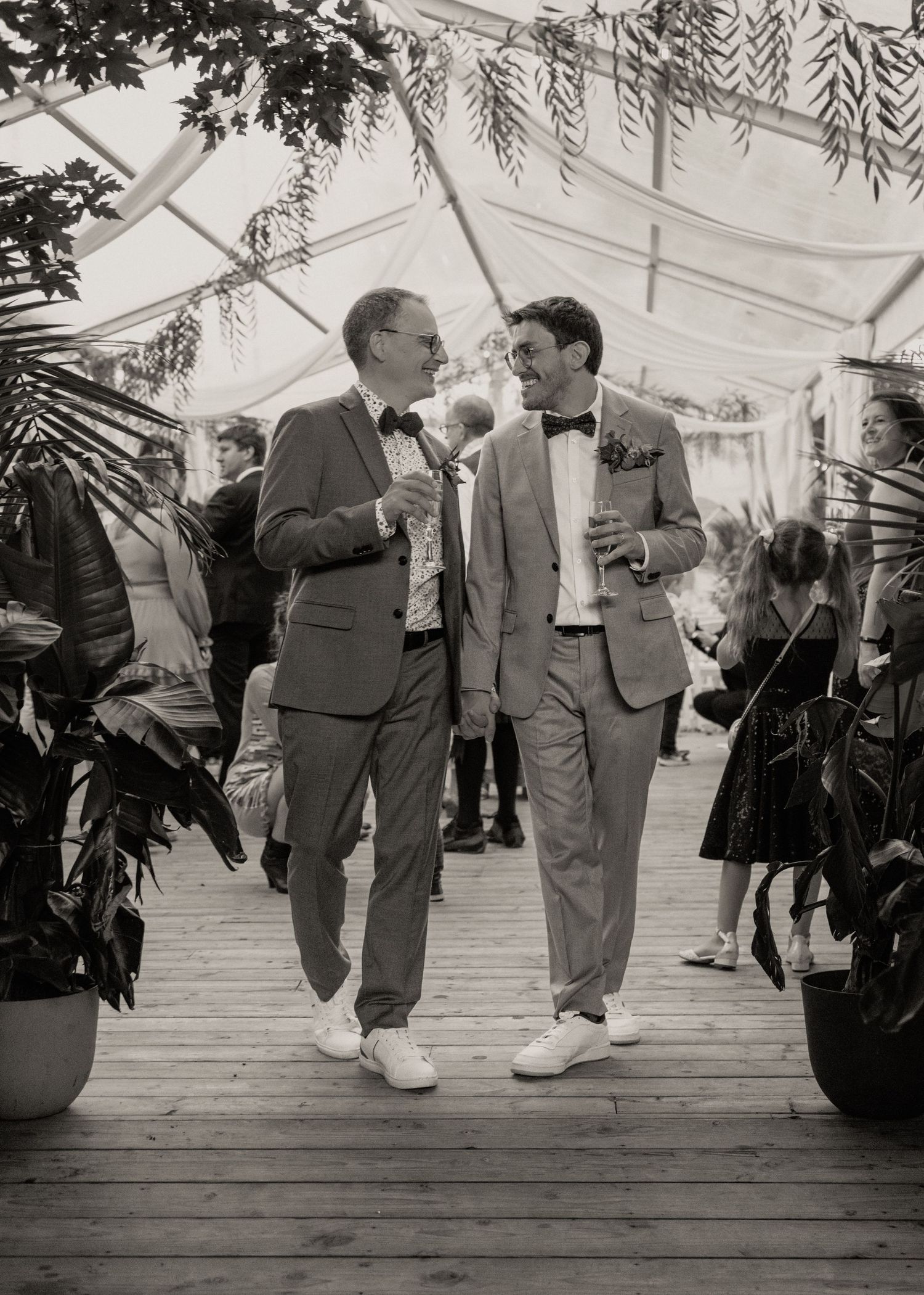 Two grooms in matching suits and bow ties share a joyful moment at their wedding reception surrounded by lush greenery.