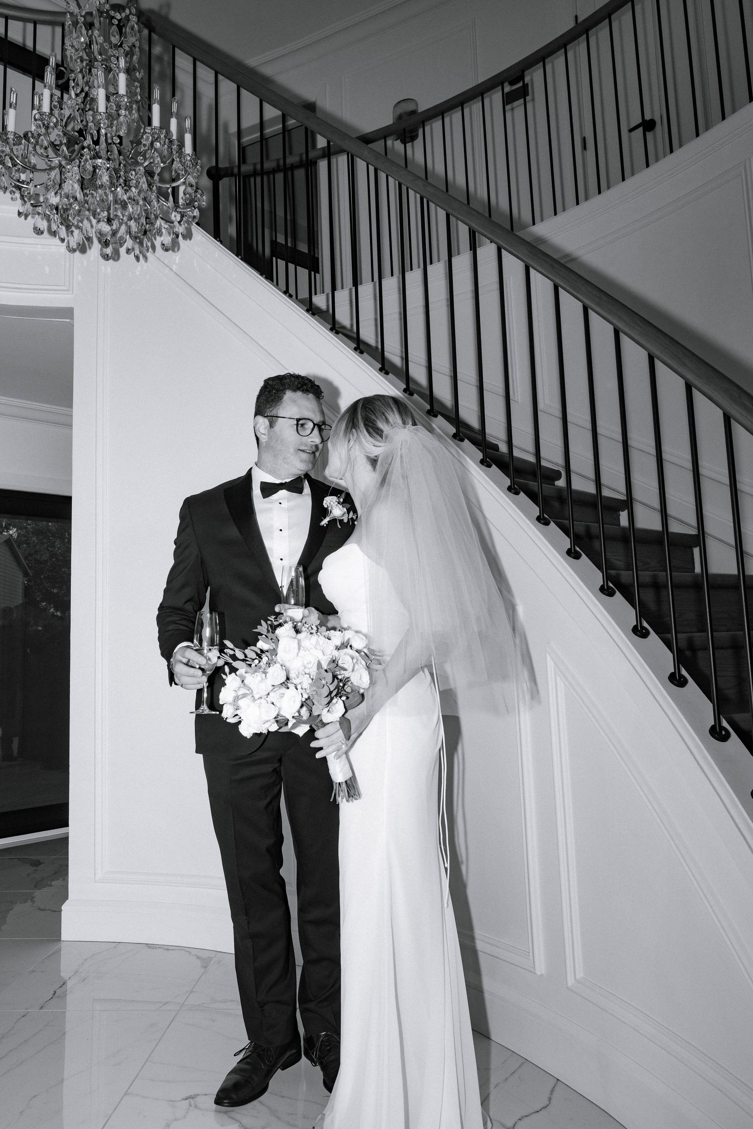 Black and white photo of bride and groom sharing intimate moment on grand staircase with crystal chandelier.