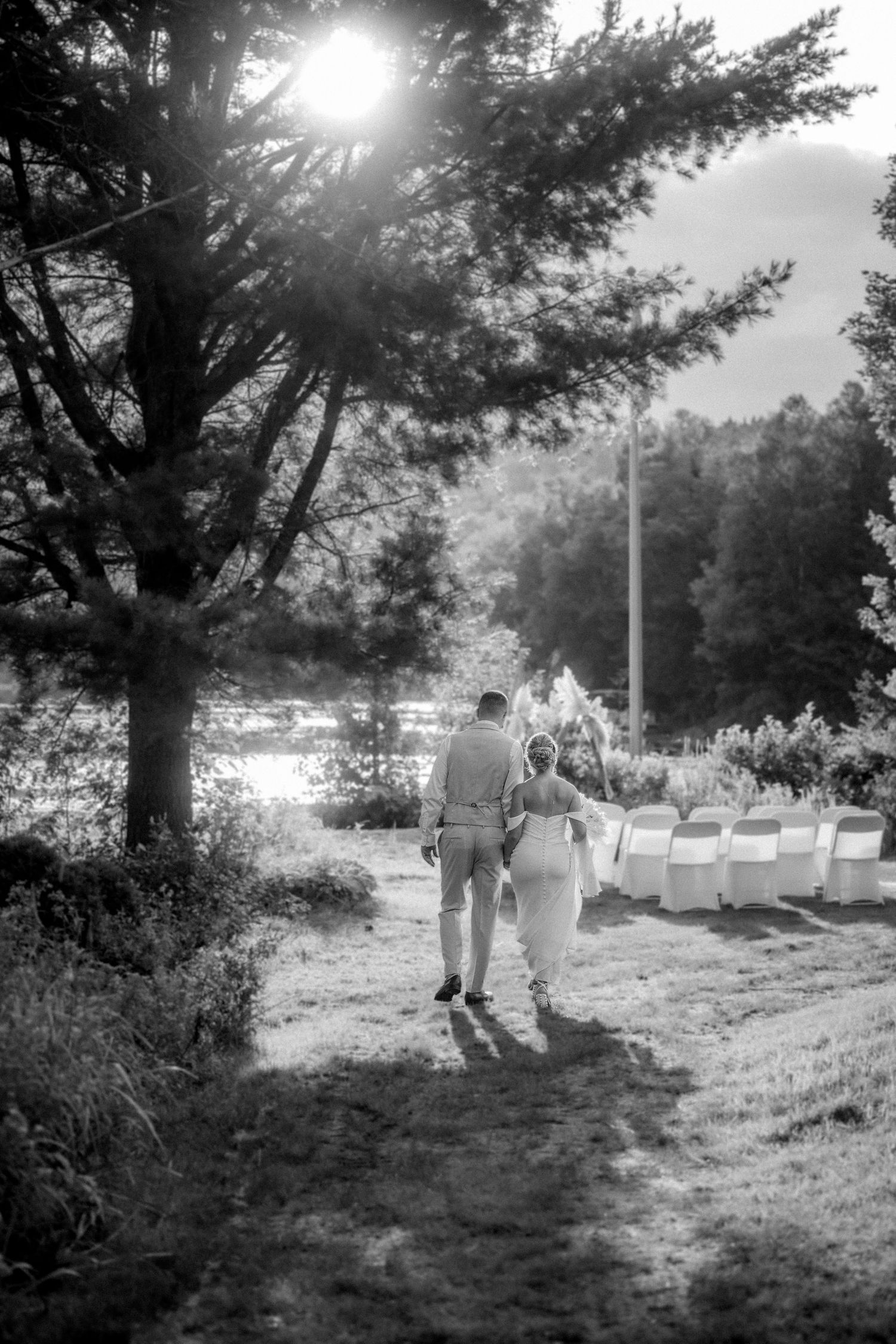 Black and white image of newlyweds strolling past rows of white chairs at an outdoor lakeside wedding ceremony.