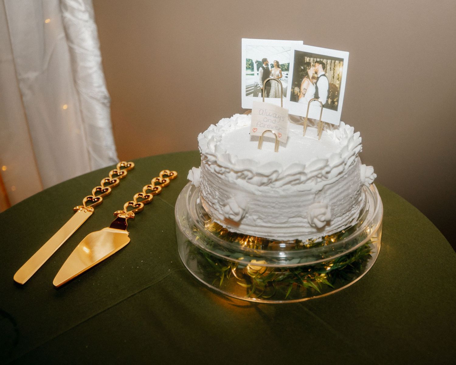 Elegant white wedding cake topped with Polaroid photos and gold cake serving set on green tablecloth.