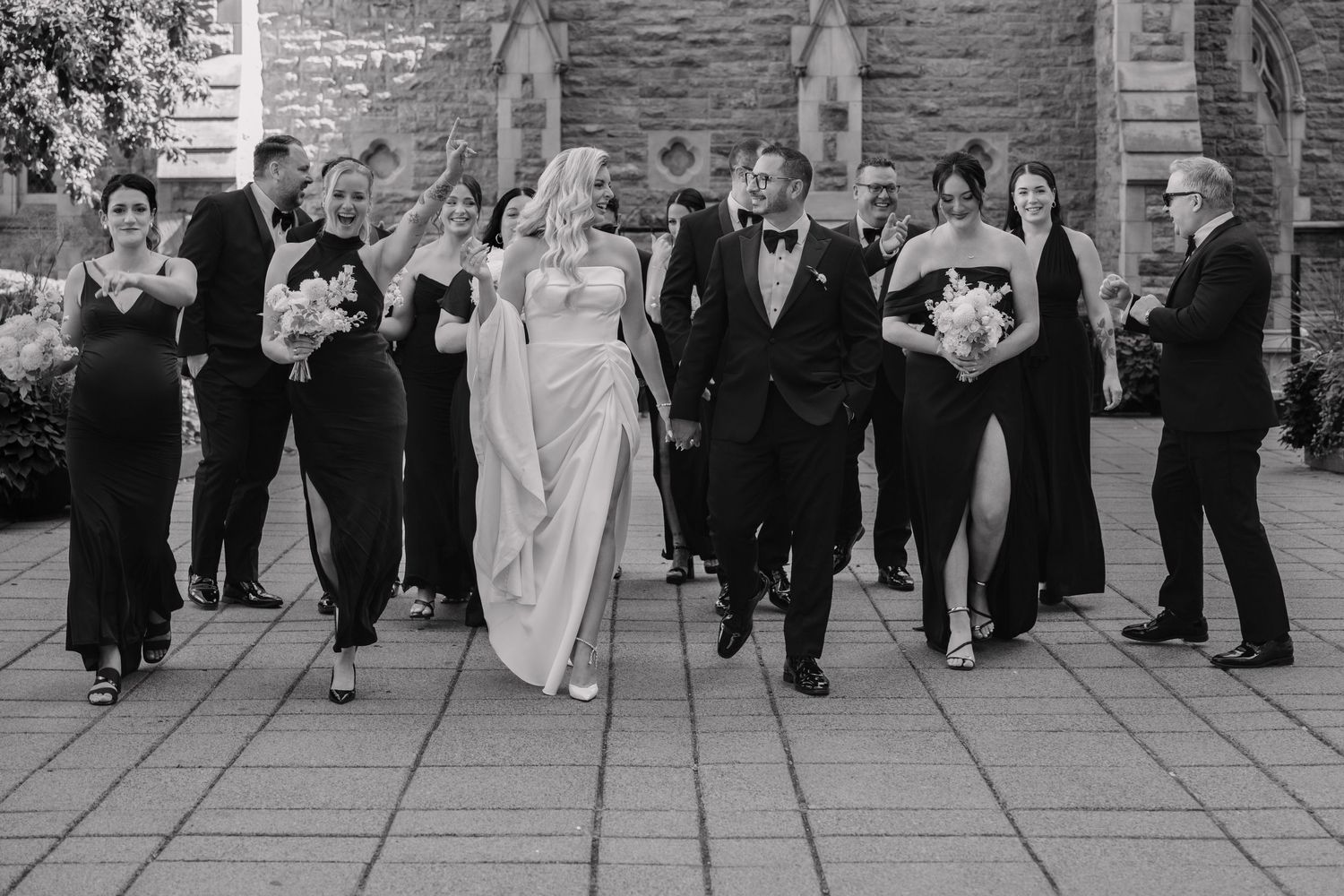 Black and white photo of a bride and groom walking with wedding party outside a stone church.