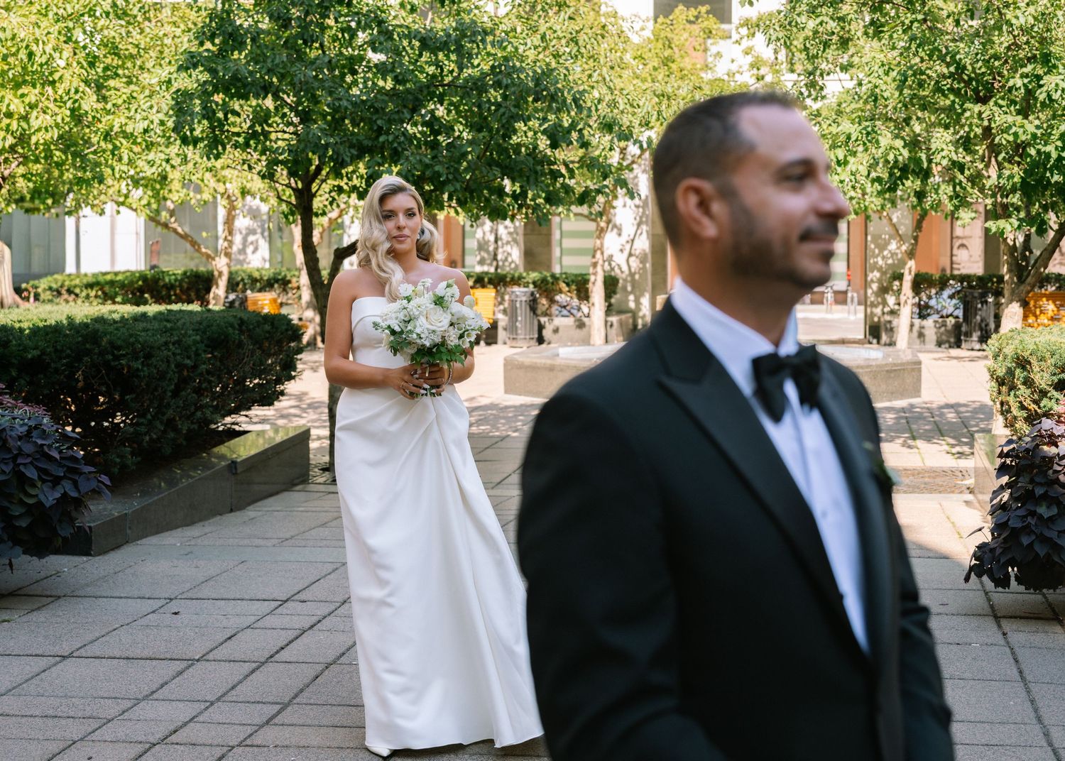 Bride in elegant white gown holds bouquet as groom in tuxedo awaits their first look outdoors.