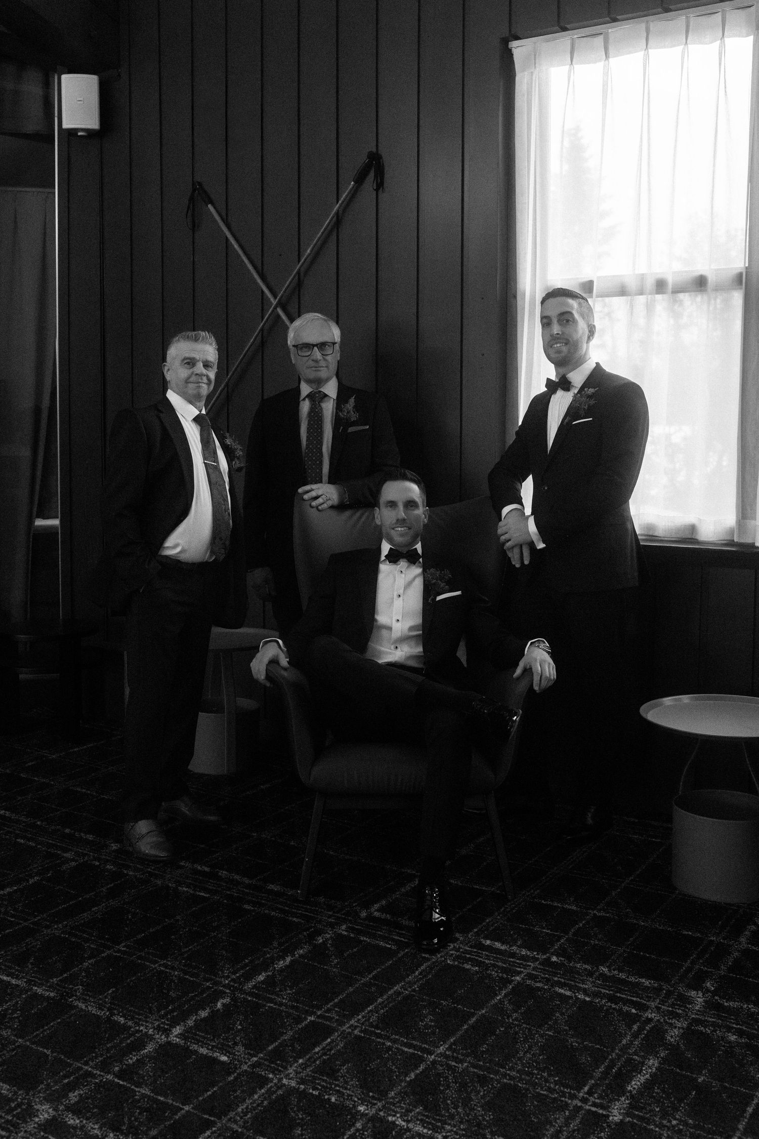 Black and white photo of four groomsmen in tuxedos posing in elegant dark-paneled room.