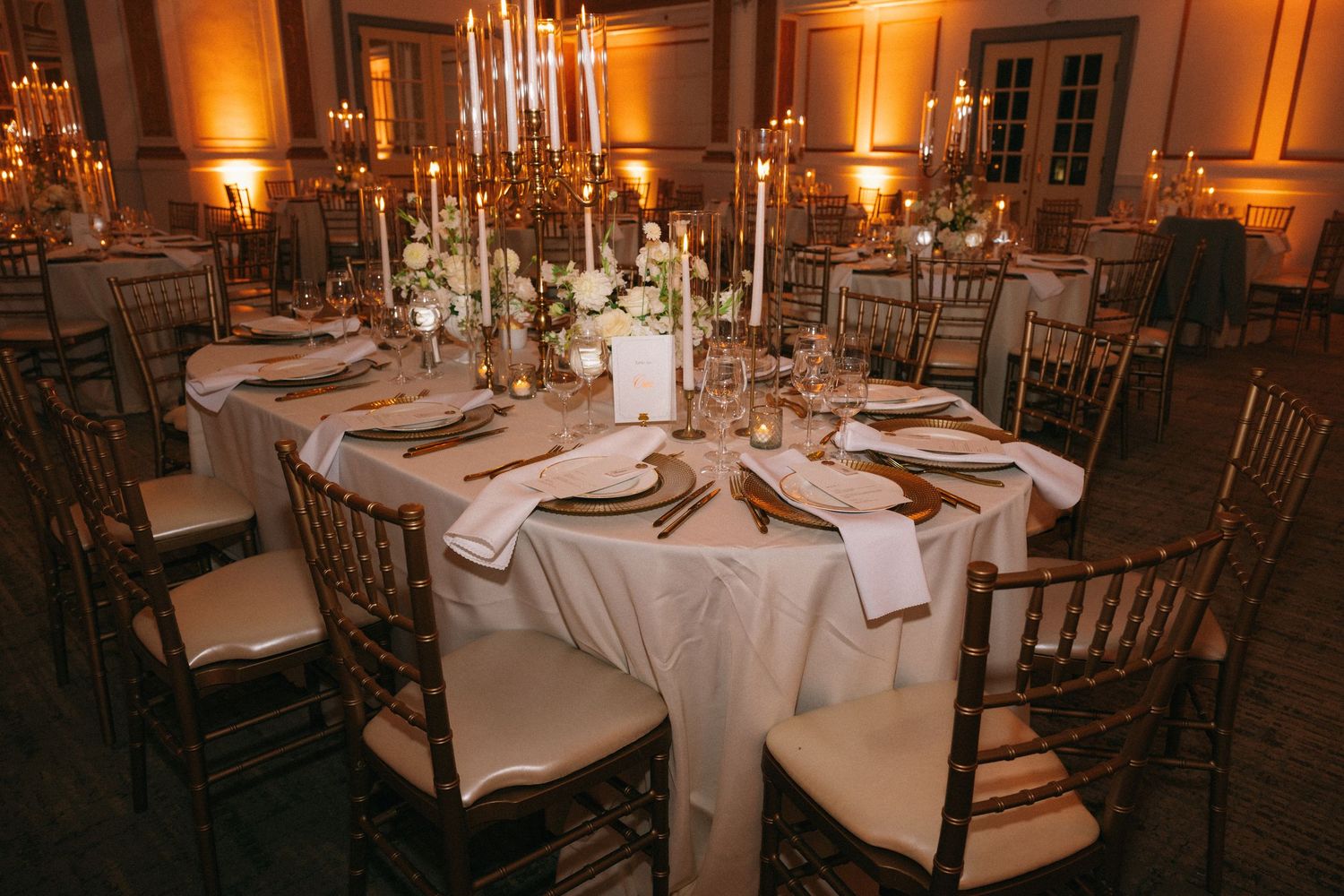 Elegant wedding reception table with gold Chiavari chairs, white linens, tall candelabras, and floral centerpieces in a warmly lit ballroom.