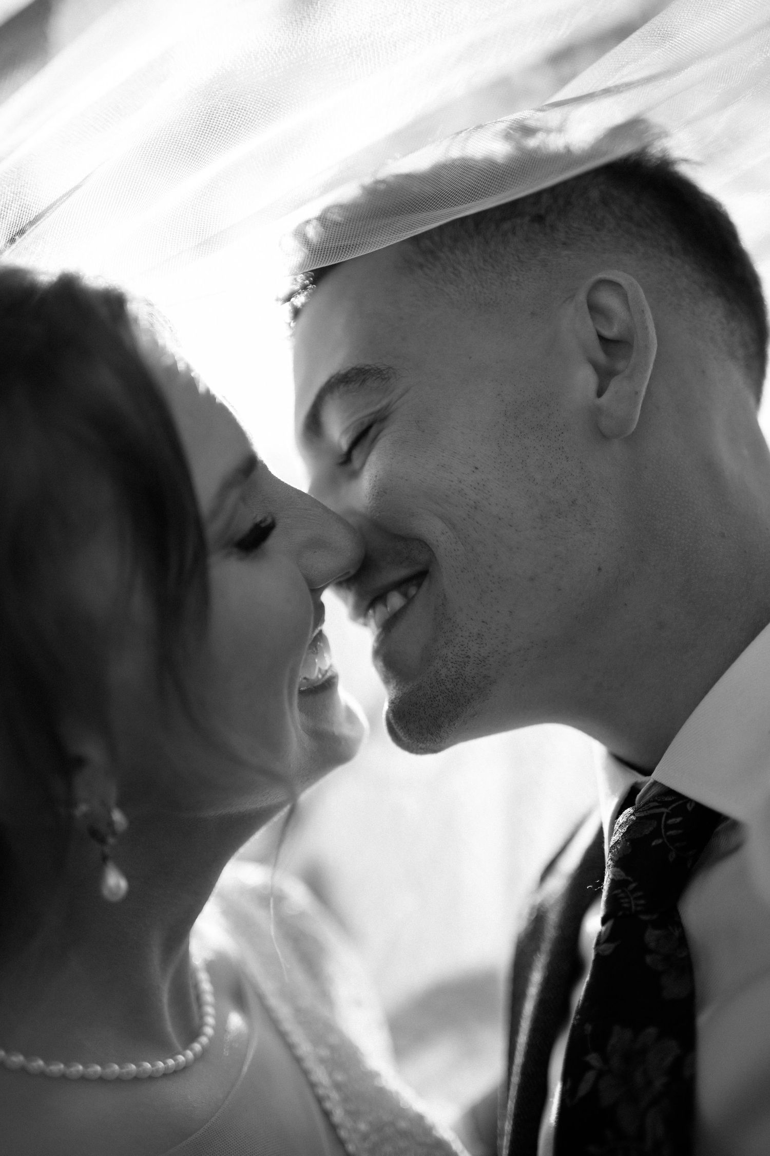 Intimate black and white close-up of laughing bride and groom under wedding veil.