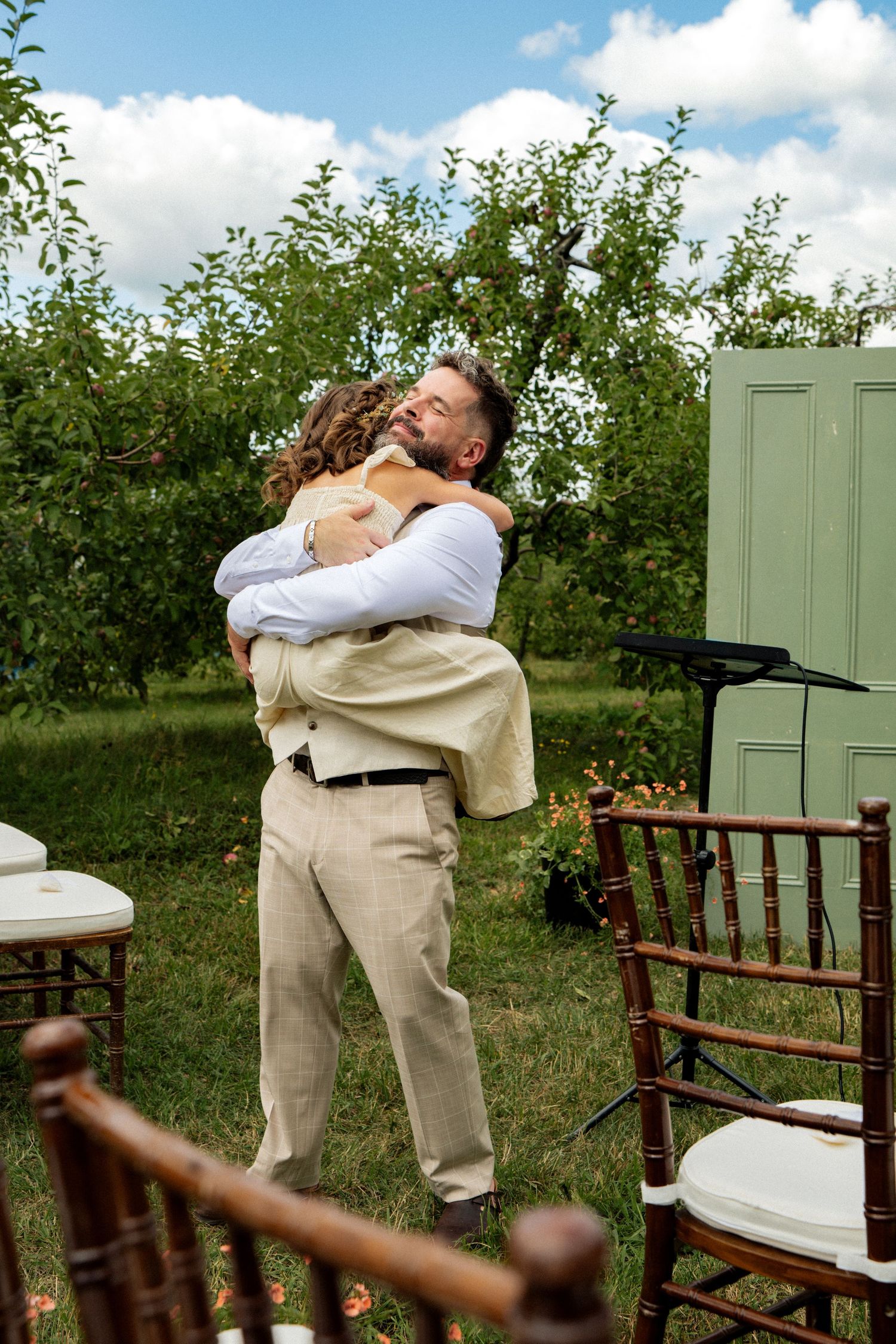 Bearded groom in linen suit lifts and embraces a child at an outdoor garden wedding ceremony.