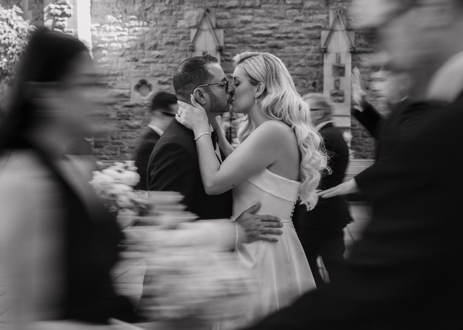 Black and white photo of bride and groom sharing a kiss surrounded by wedding guests indoors.