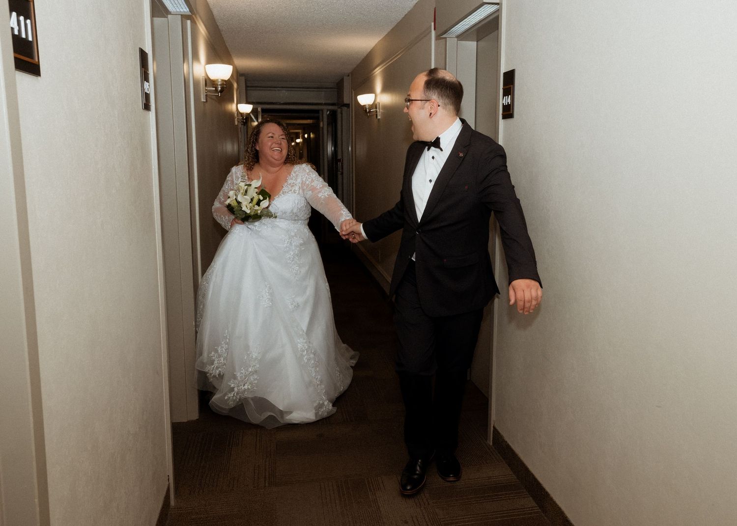 Bride in lace gown and groom in tuxedo walking hand in hand down a hotel hallway.