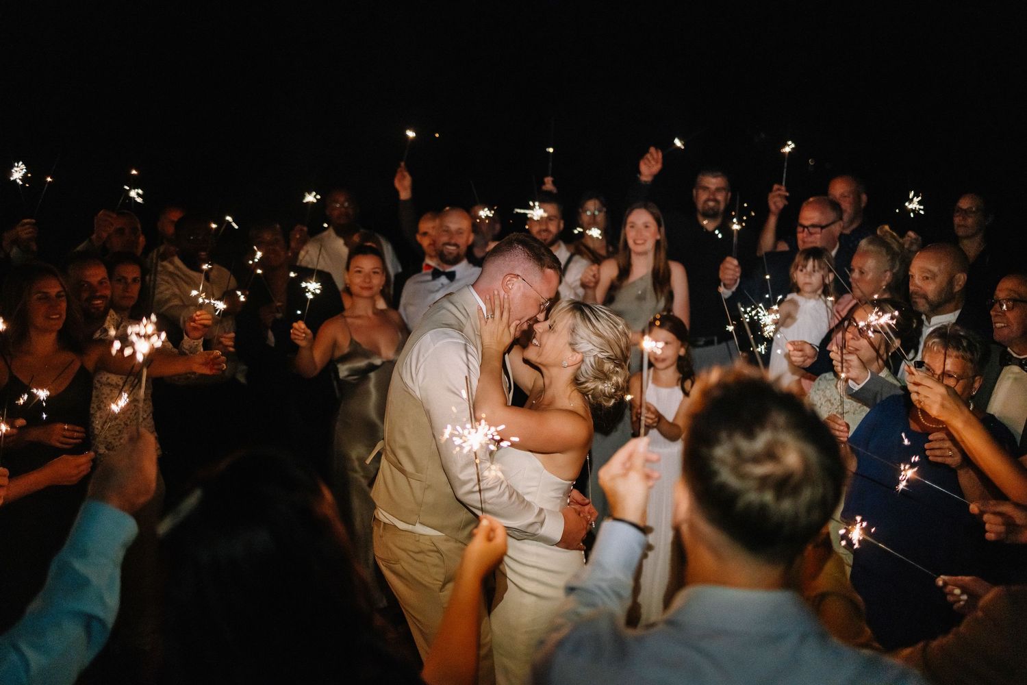 Bride and groom kiss during magical sparkler exit surrounded by celebrating wedding guests at night.