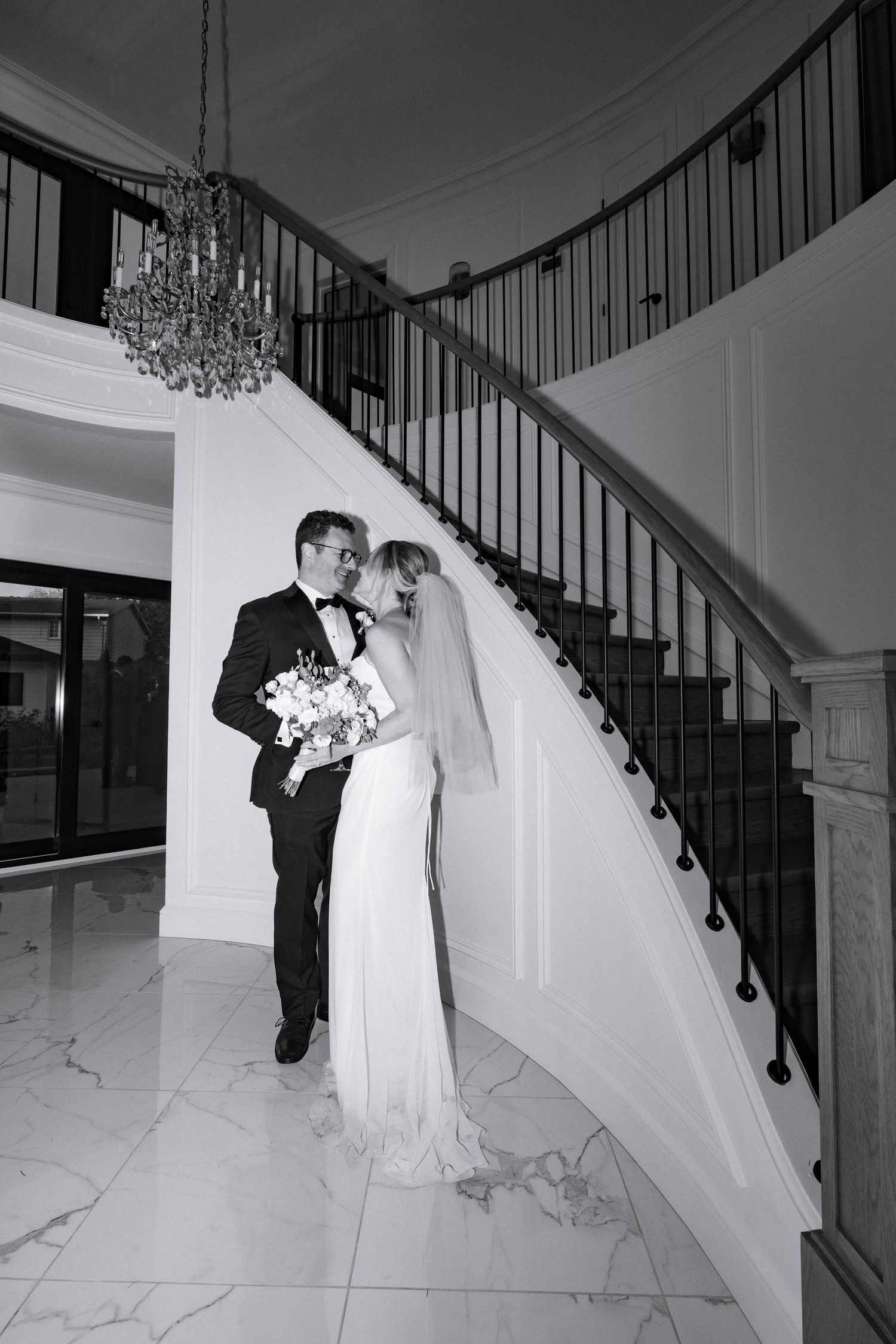 Bride and groom share romantic moment by elegant curved staircase in grand marble foyer with chandelier.